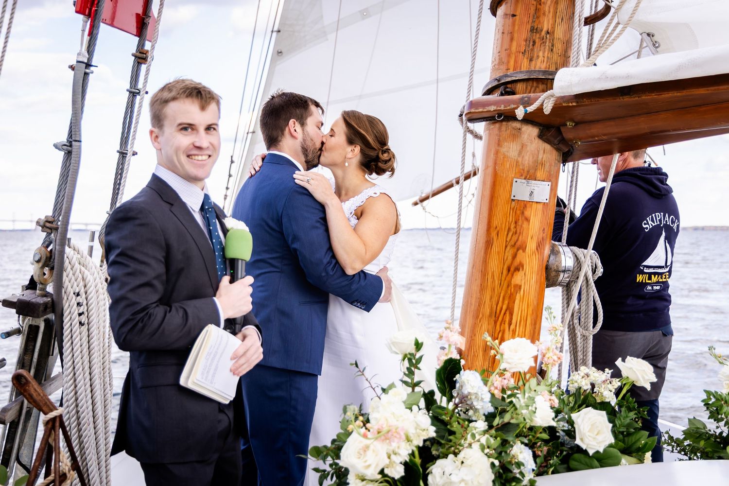 Couple kissing during outdoor sailboat wedding ceremony with officiant holding microphone and floral decorations on deck.