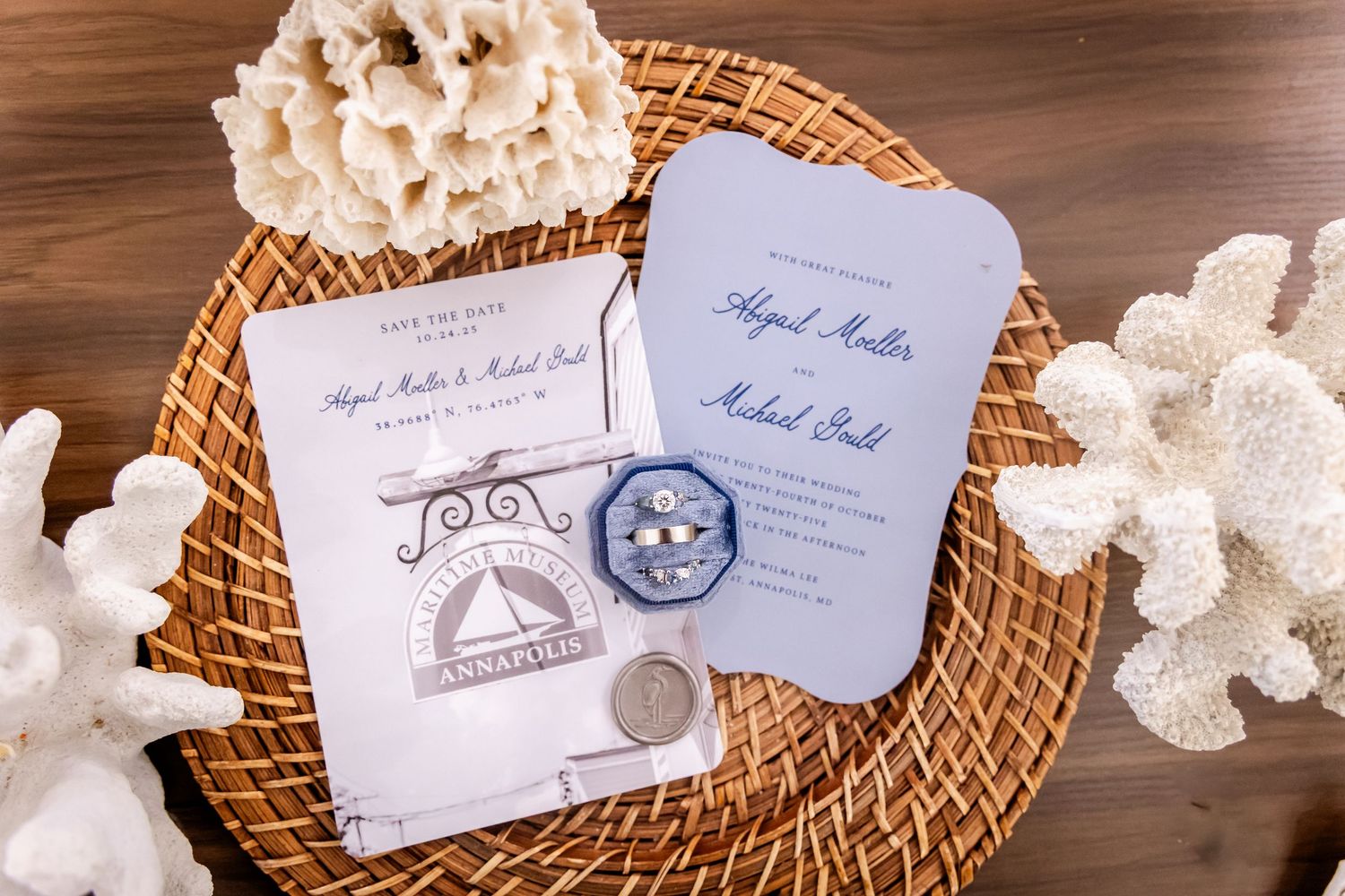 Wedding stationery flat lay with blue invitations, save the dates, rings, and coral accents on wicker tray.