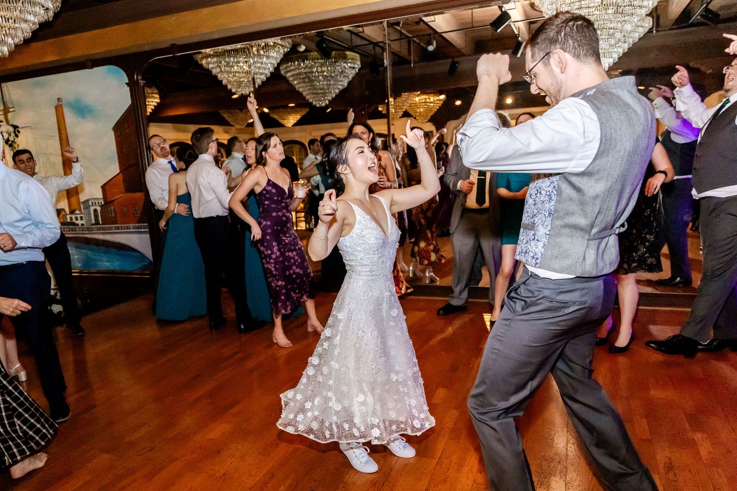 Wedding guests dancing and celebrating on a wooden dance floor at an indoor reception venue.