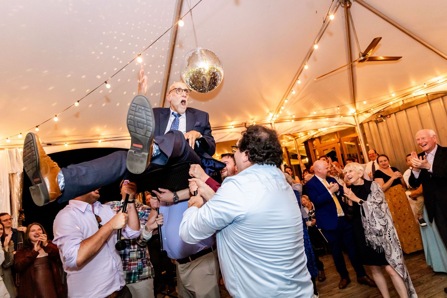 Wedding guests dancing and celebrating under a white tent with string lights and decorative lanterns during evening reception.