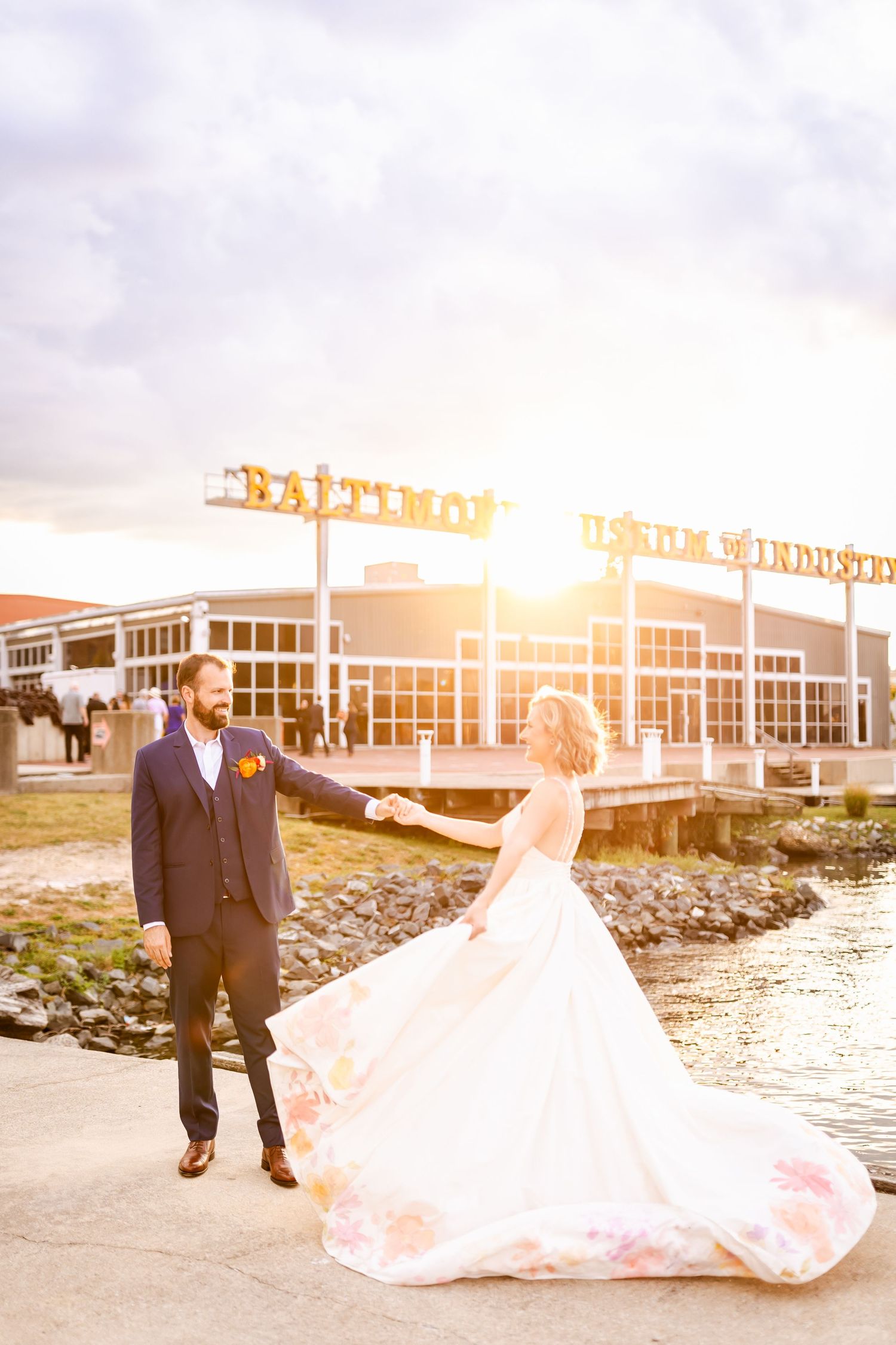 Newlywed couple poses along rocky shoreline at sunset with wedding dress flowing in the breeze.