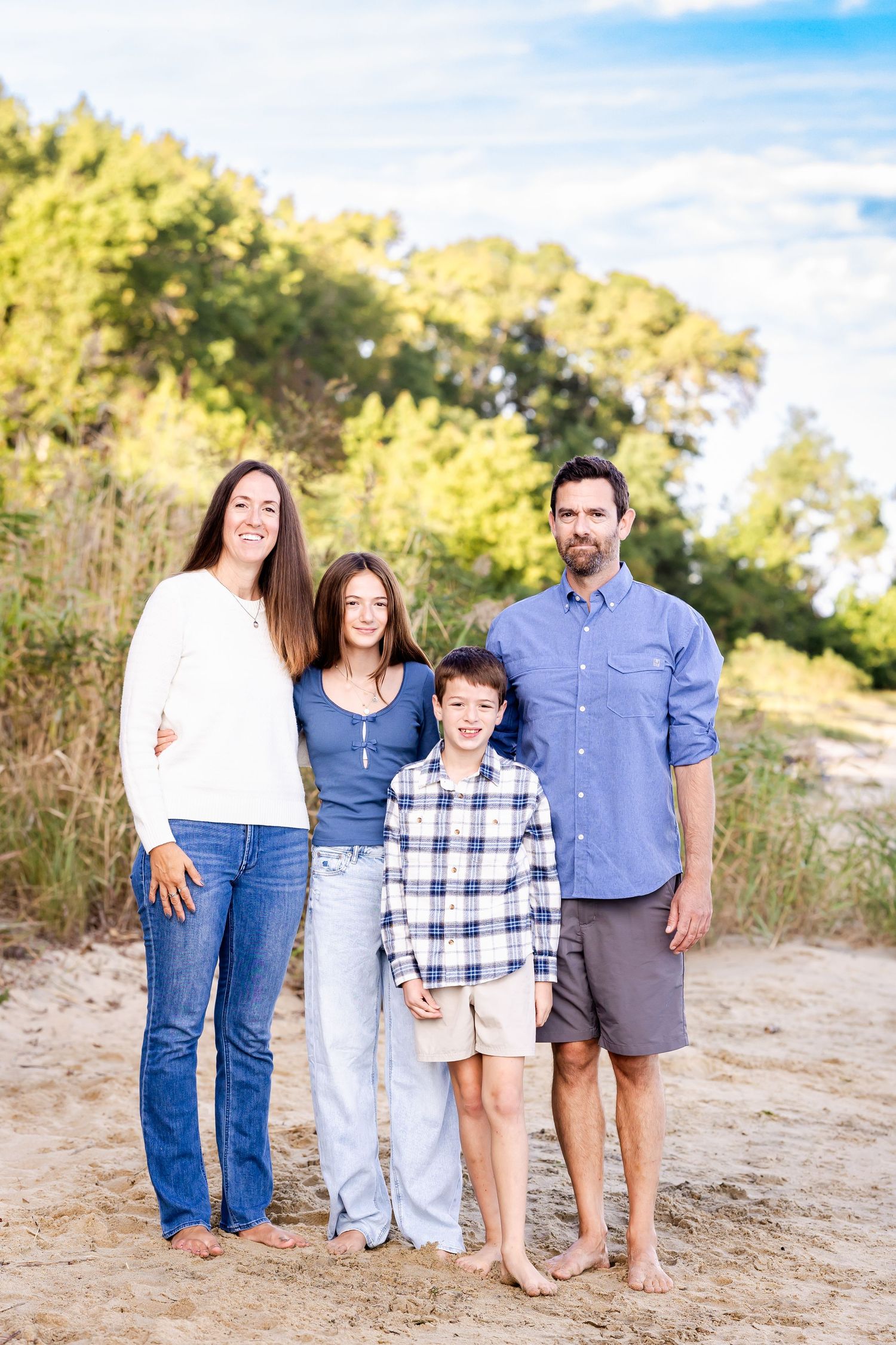 Casual family portrait session on a sandy beach with blue skies and greenery.