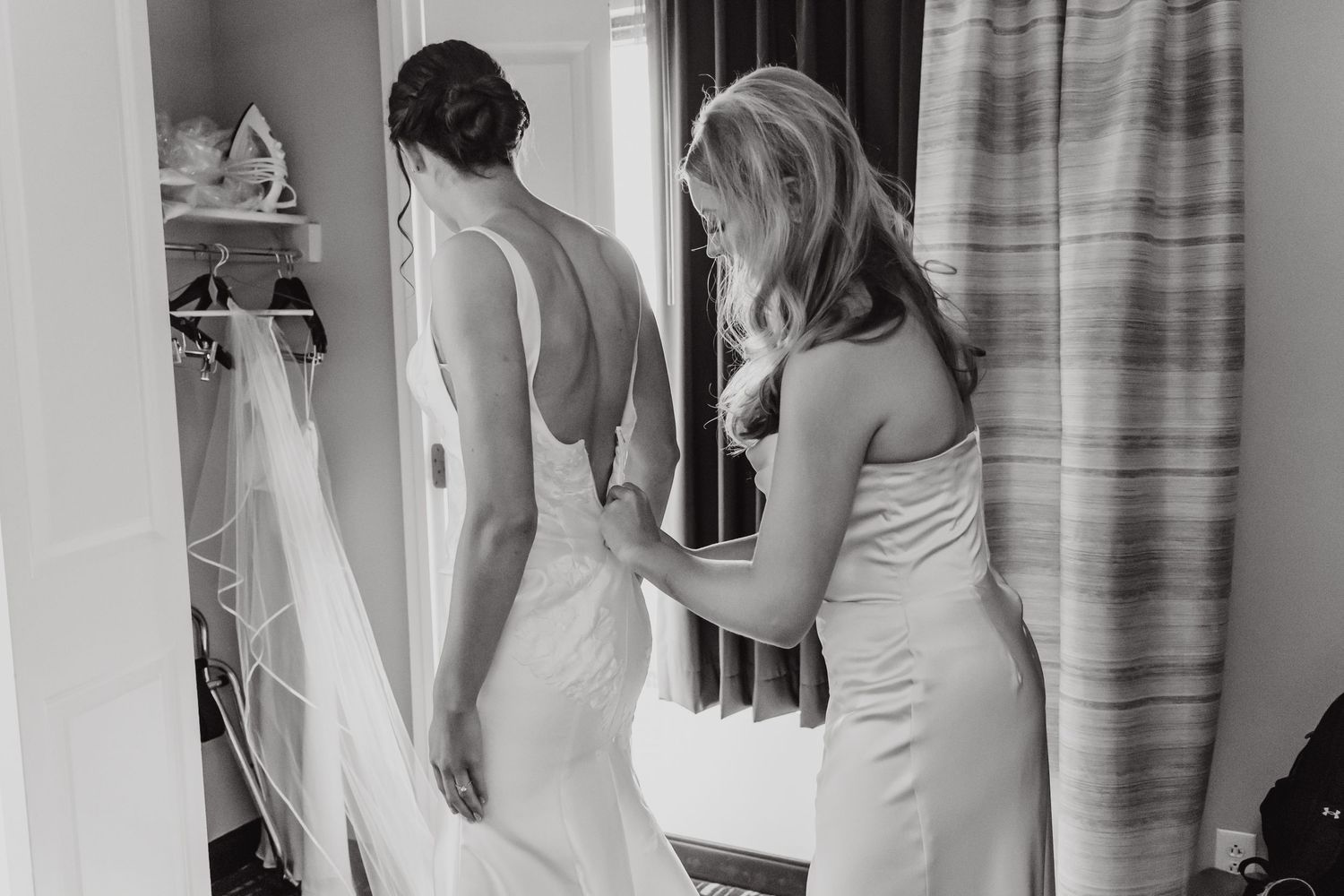 A black and white photo of a bride getting help with her wedding dress during preparation.
