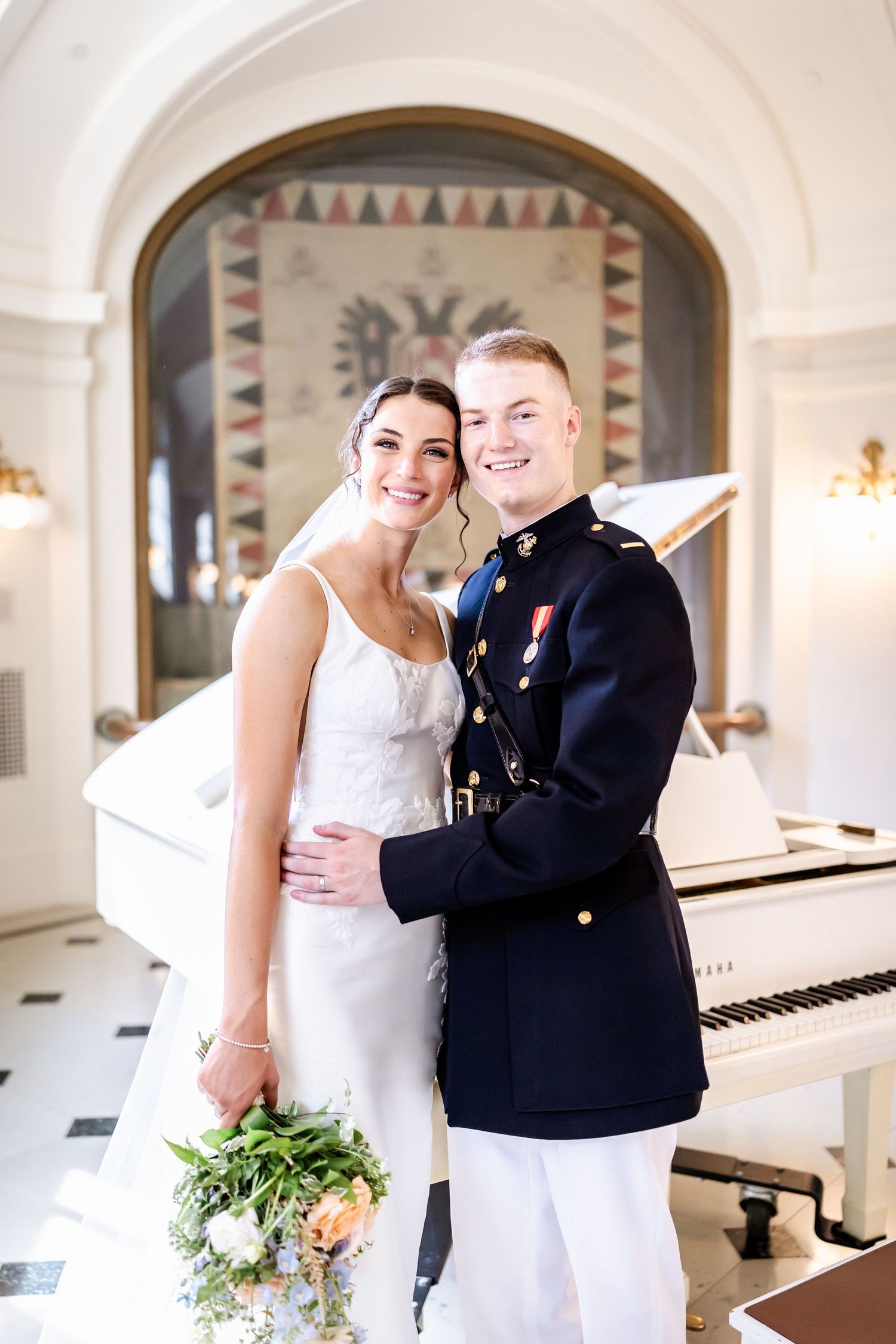 Military wedding couple sharing joyful moments in an elegant indoor venue with white decor and piano.