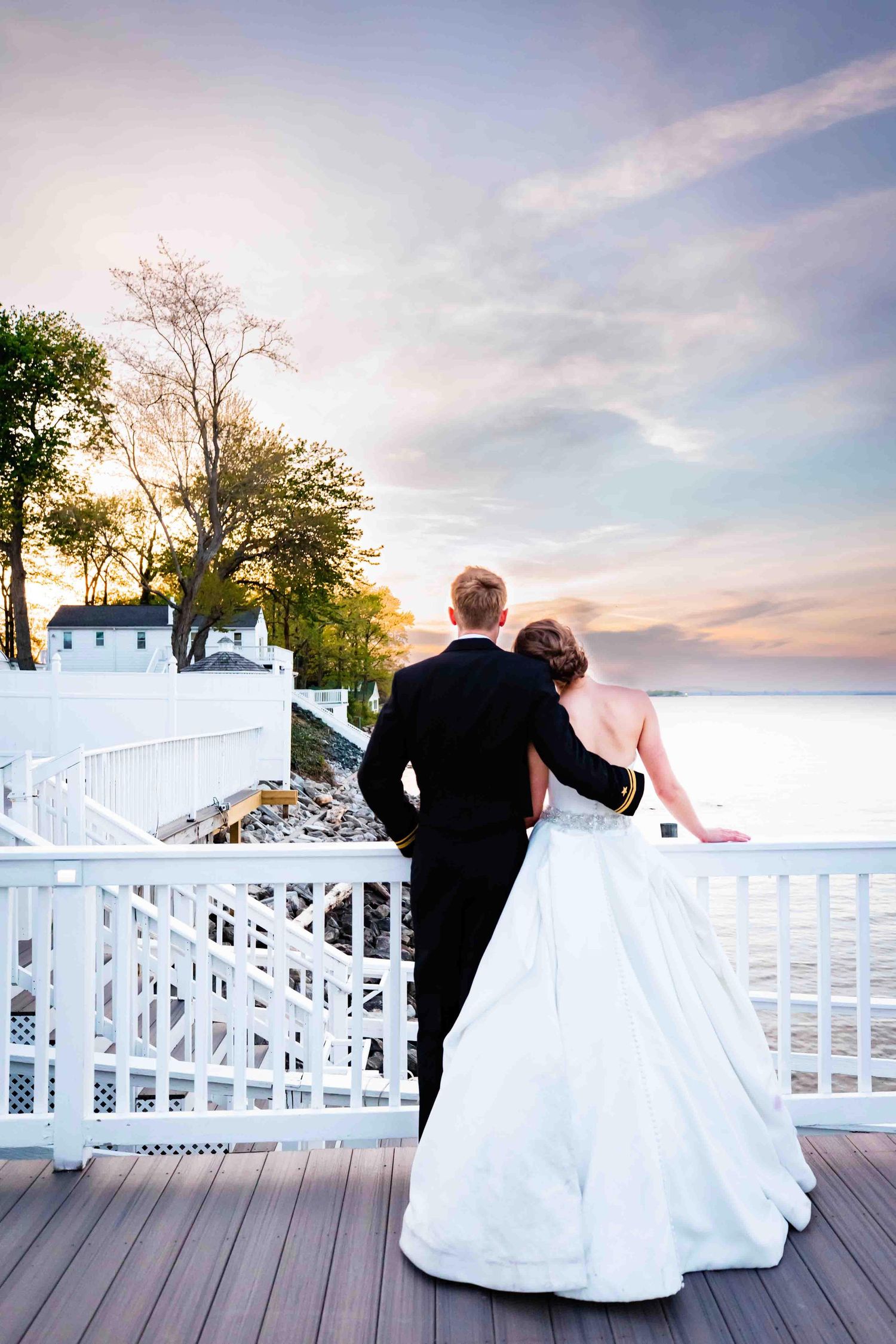 A couple in formal wedding attire stands on a white deck overlooking a scenic waterfront at sunset.