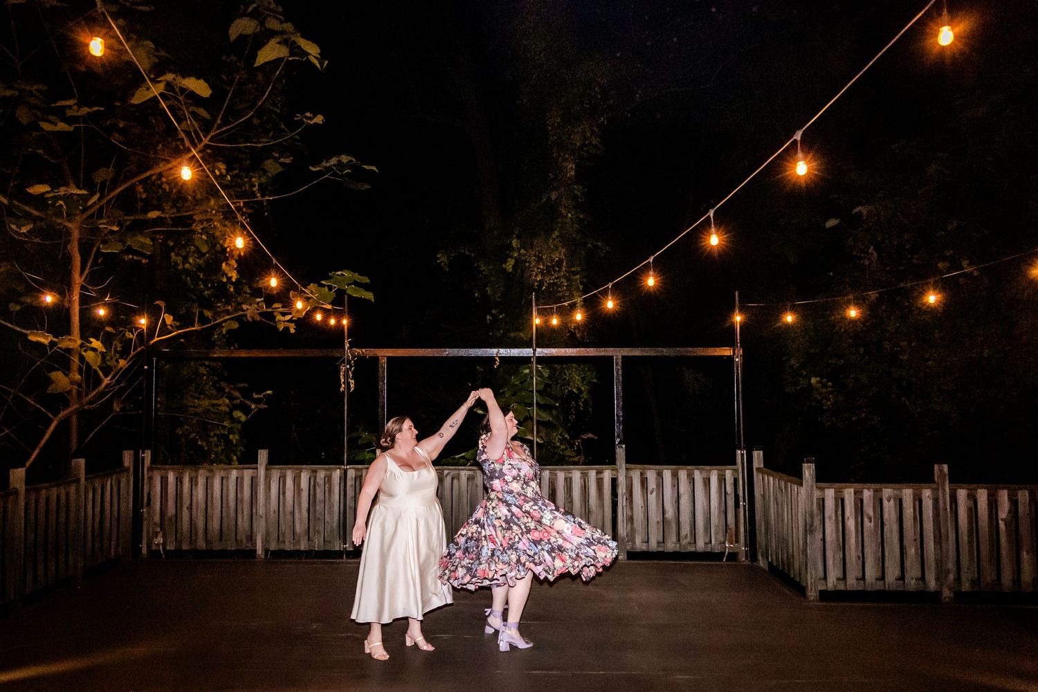 Two dancers twirl beneath glowing string lights at an outdoor evening celebration.