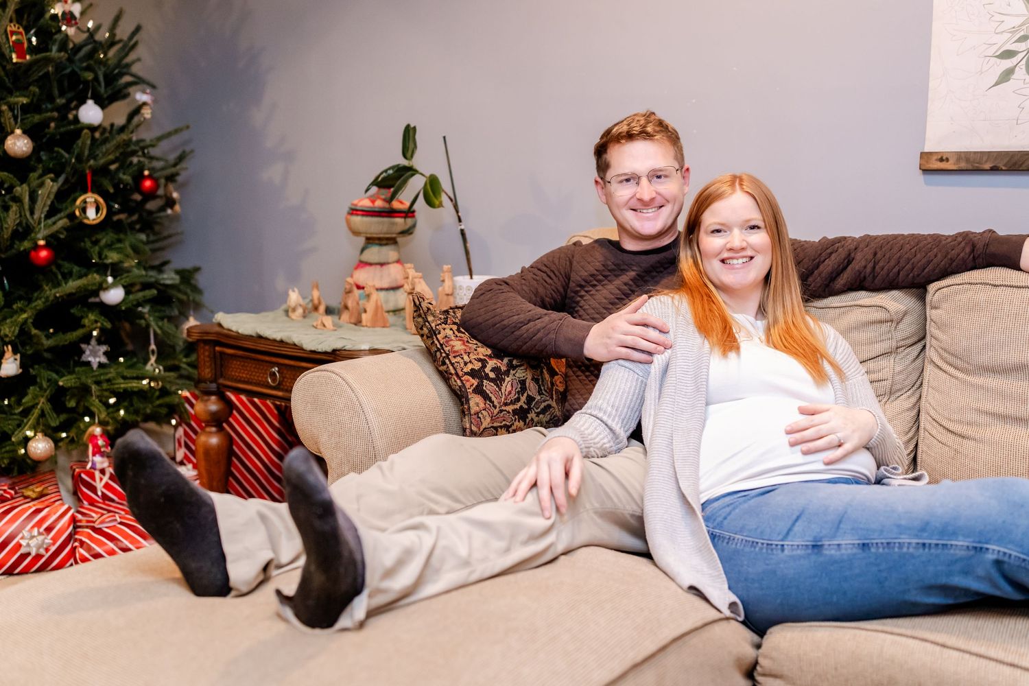 A couple relaxes on a couch next to a decorated Christmas tree with wrapped presents underneath.