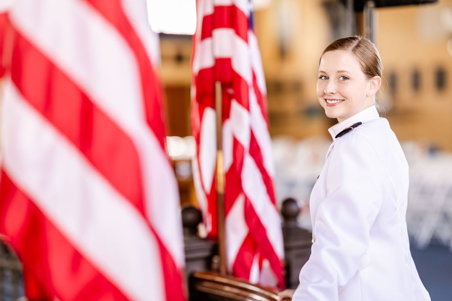 A person in a white uniform stands beside American flags in a formal setting.