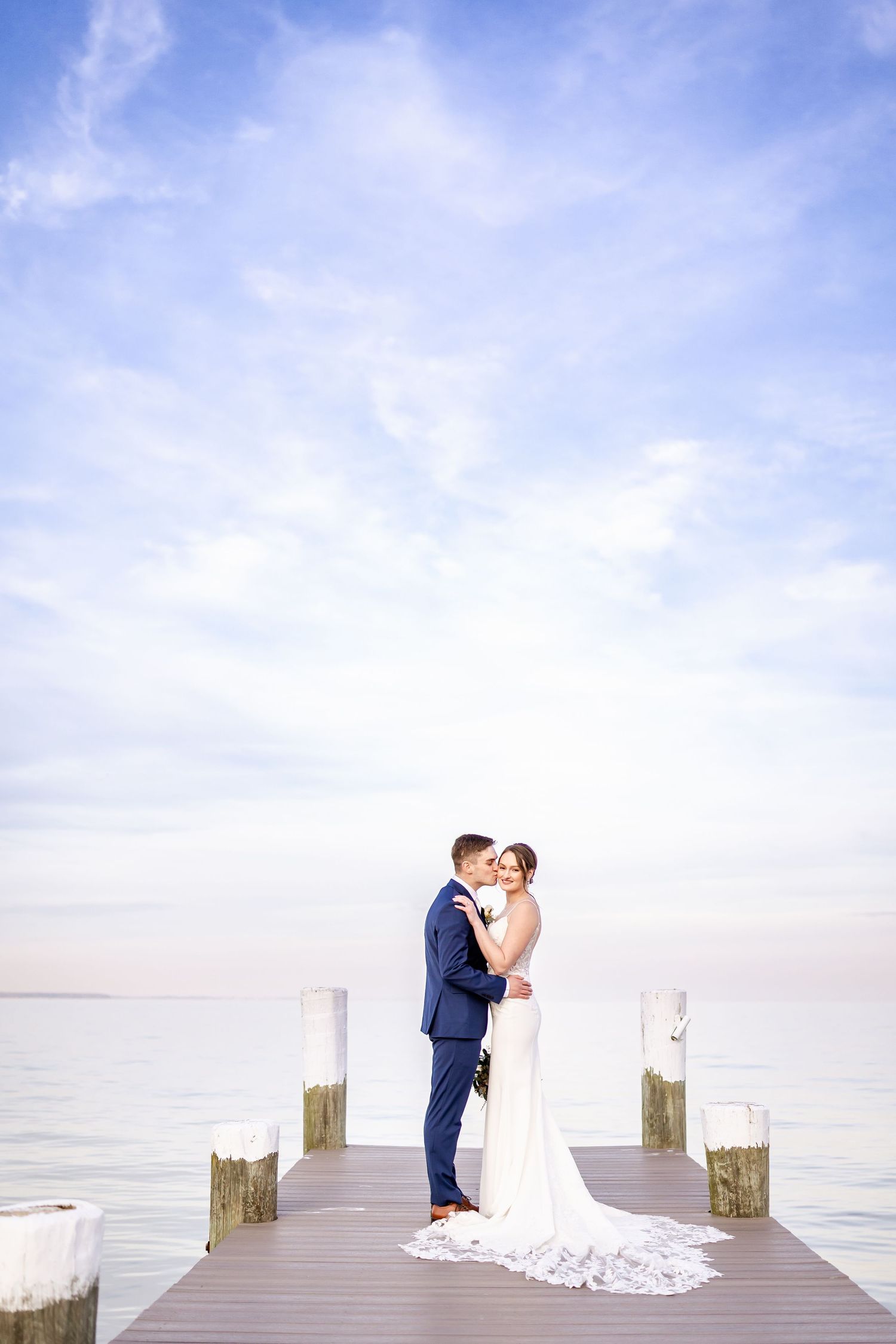Newlyweds pose for romantic portraits on a dock with bright blue sky and water backdrop.