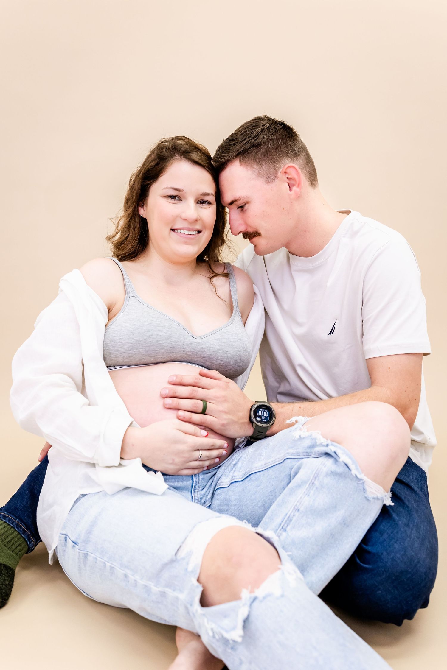 An intimate maternity photo with a couple in casual wear sitting close together against neutral backdrop.
