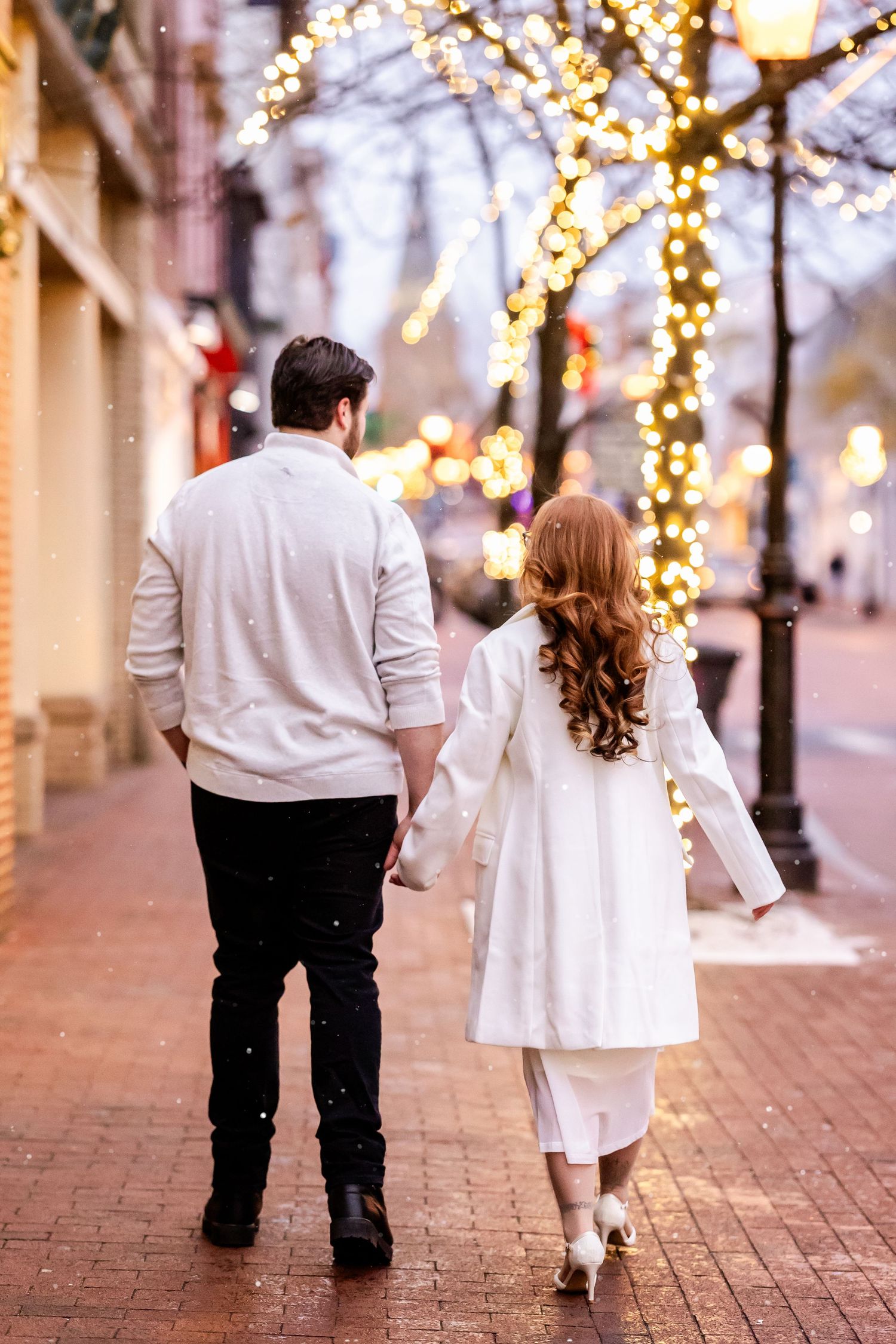 A couple walks hand in hand down a brick pathway decorated with twinkling string lights in winter.