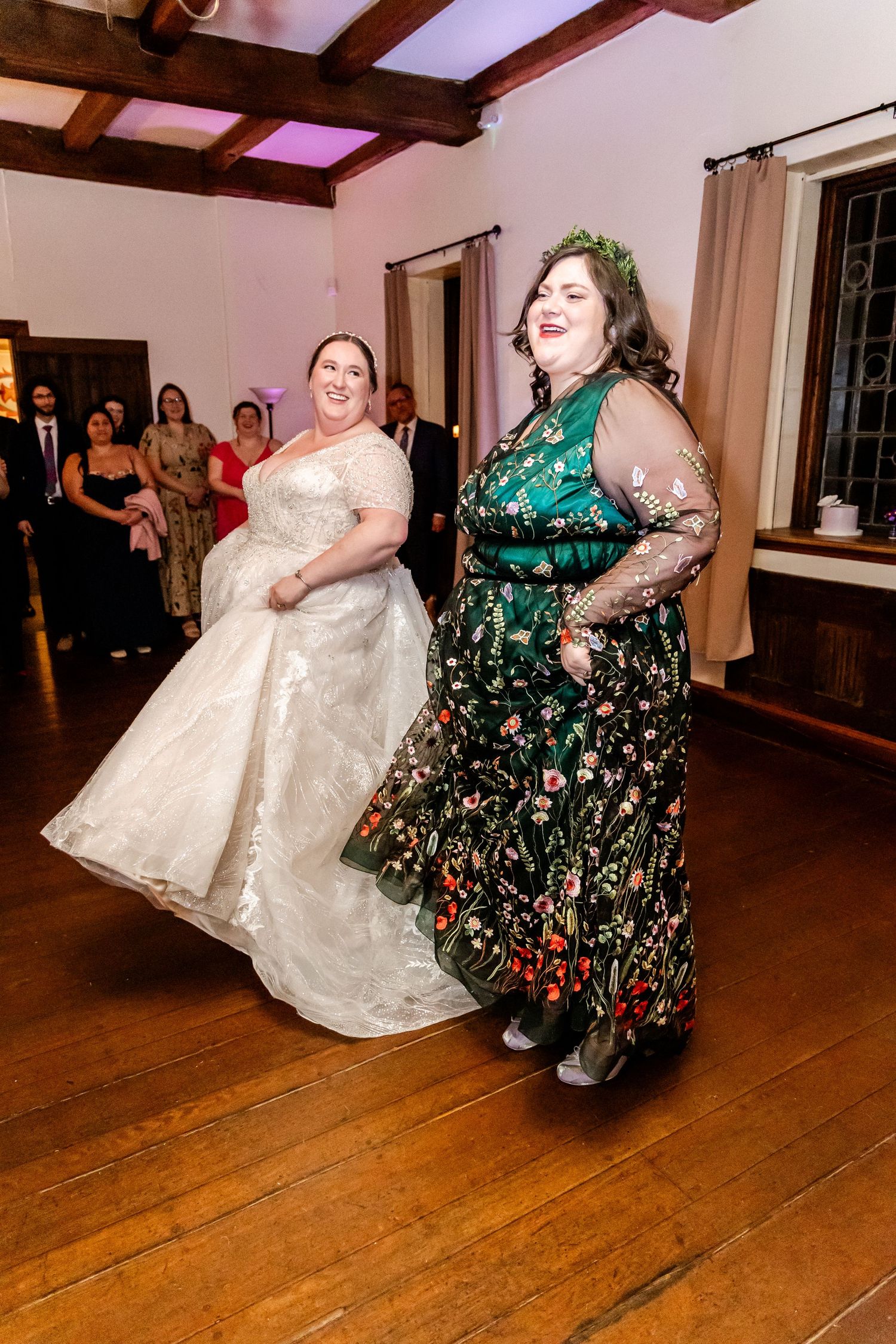 Two people in elegant dresses dancing and having fun at a wedding reception in a wooden-floored room.