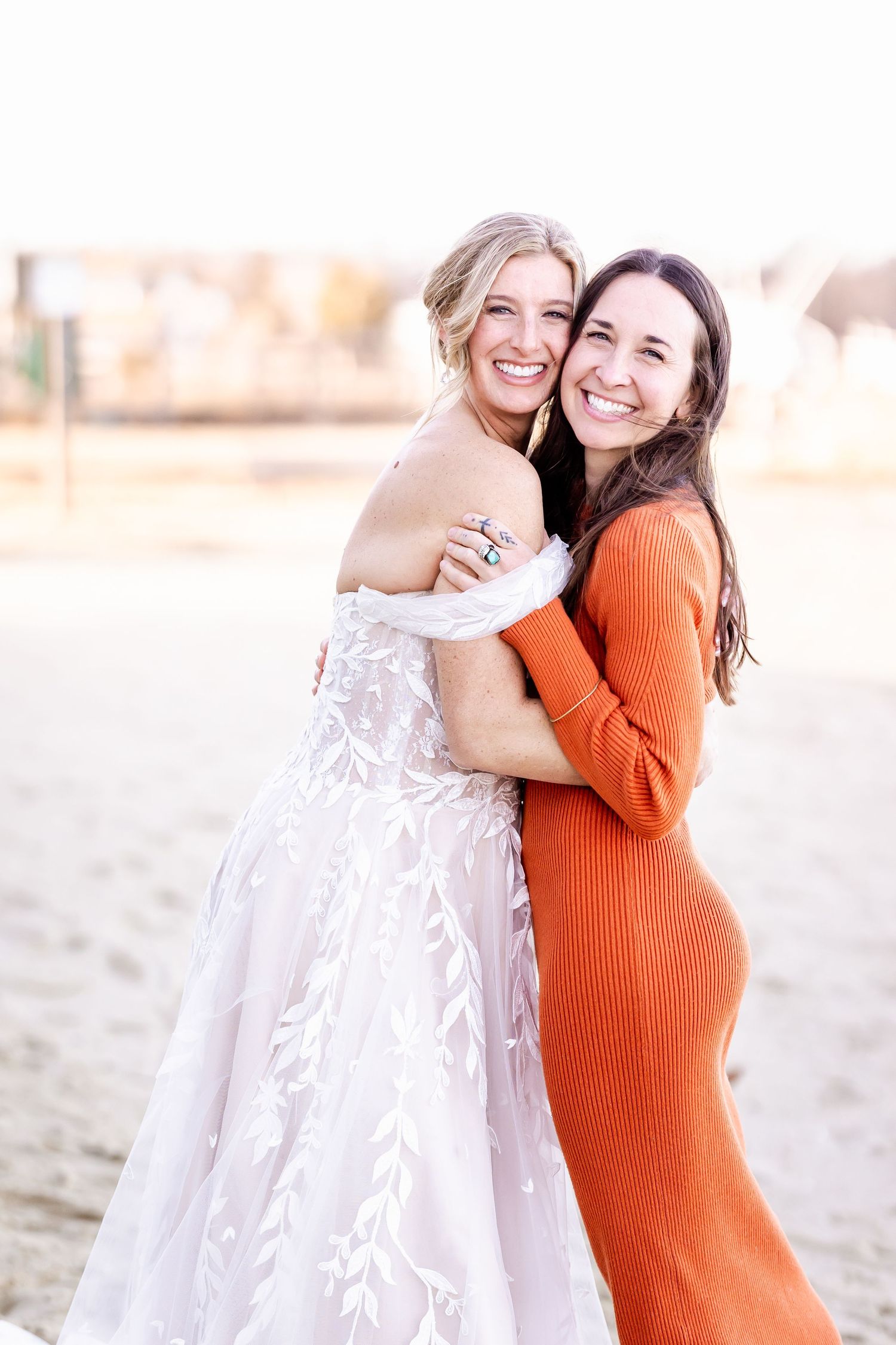 Two people hugging and smiling outdoors, wearing a white wedding gown and fitted orange dress.
