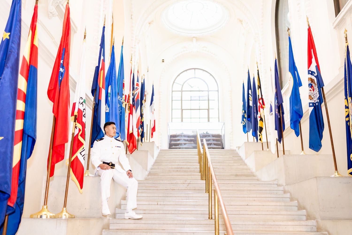 Person in white clothing stands beside colorful flags displayed along a grand marble staircase in light-filled hallway.