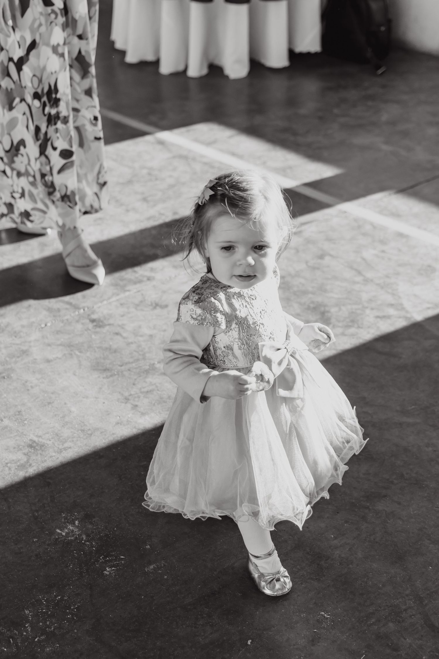 A small child wearing a white lace dress and sandals stands in a sunlit reception hall.