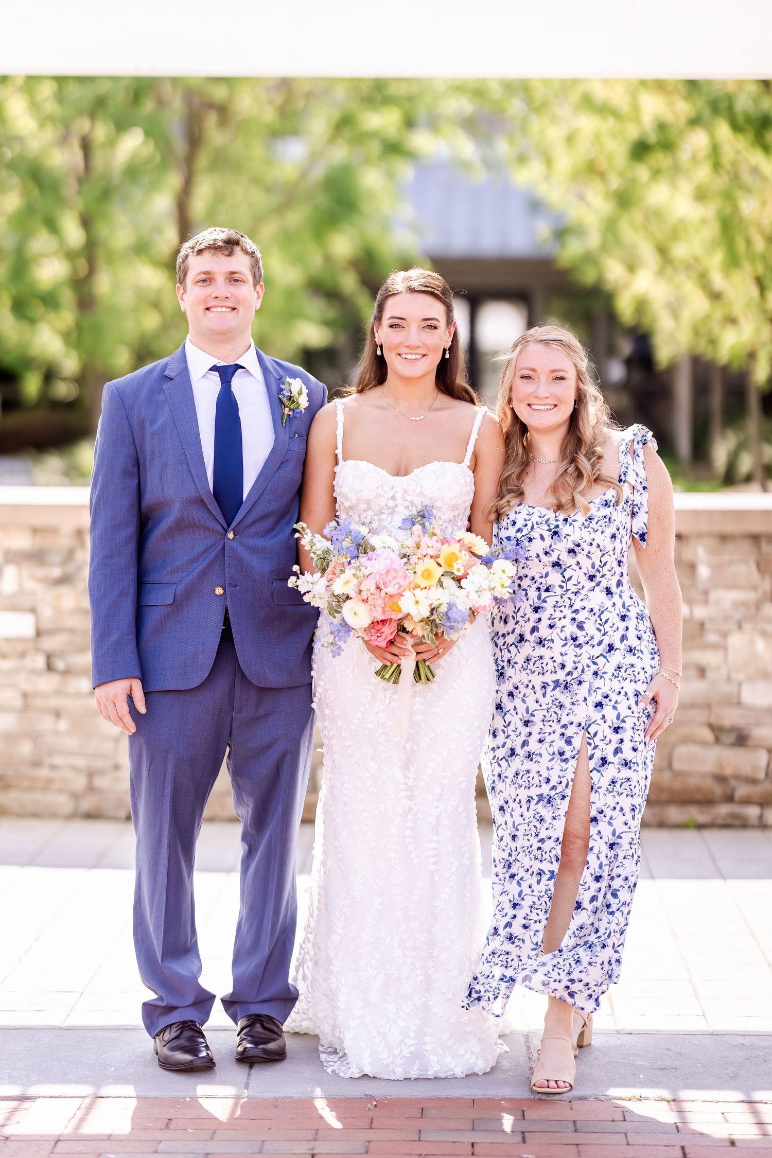 Wedding party in blue suits and floral dresses poses together outdoors on a sunny day.