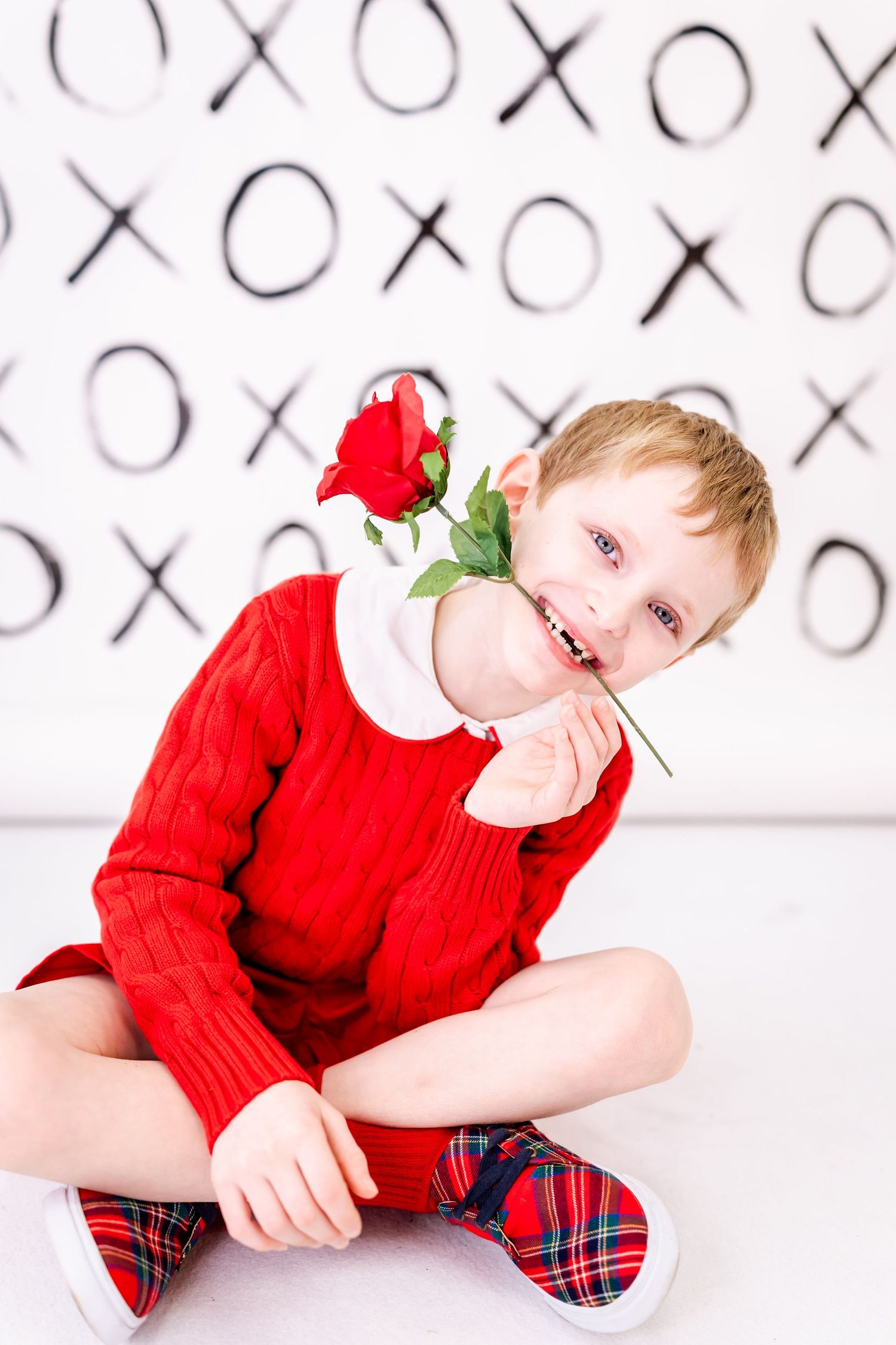 A small child in a red sweater sits cross-legged on a white floor holding a red rose against a pattern background.