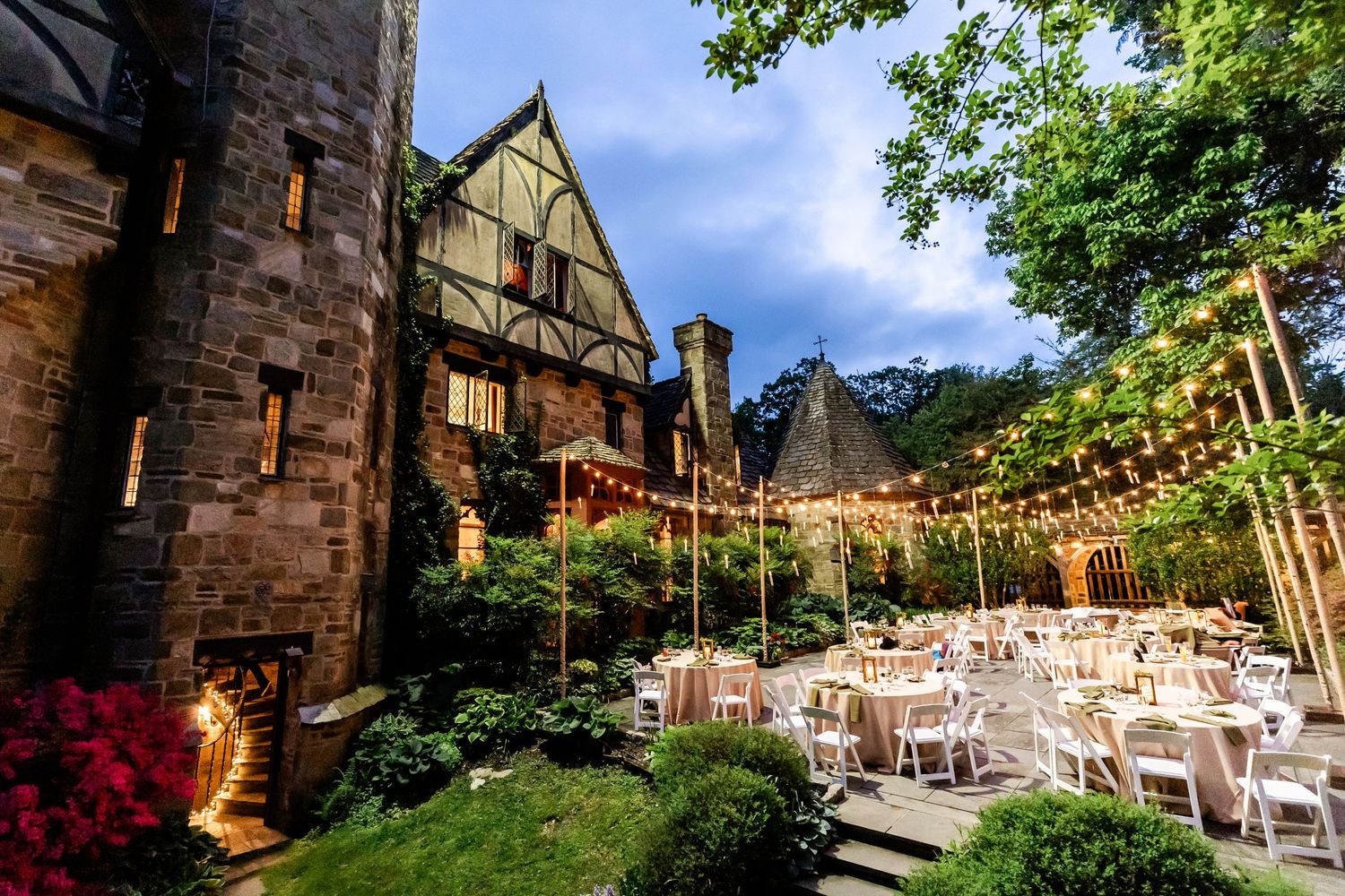 Gothic stone mansion decorated for outdoor wedding reception with string lights and white tables.