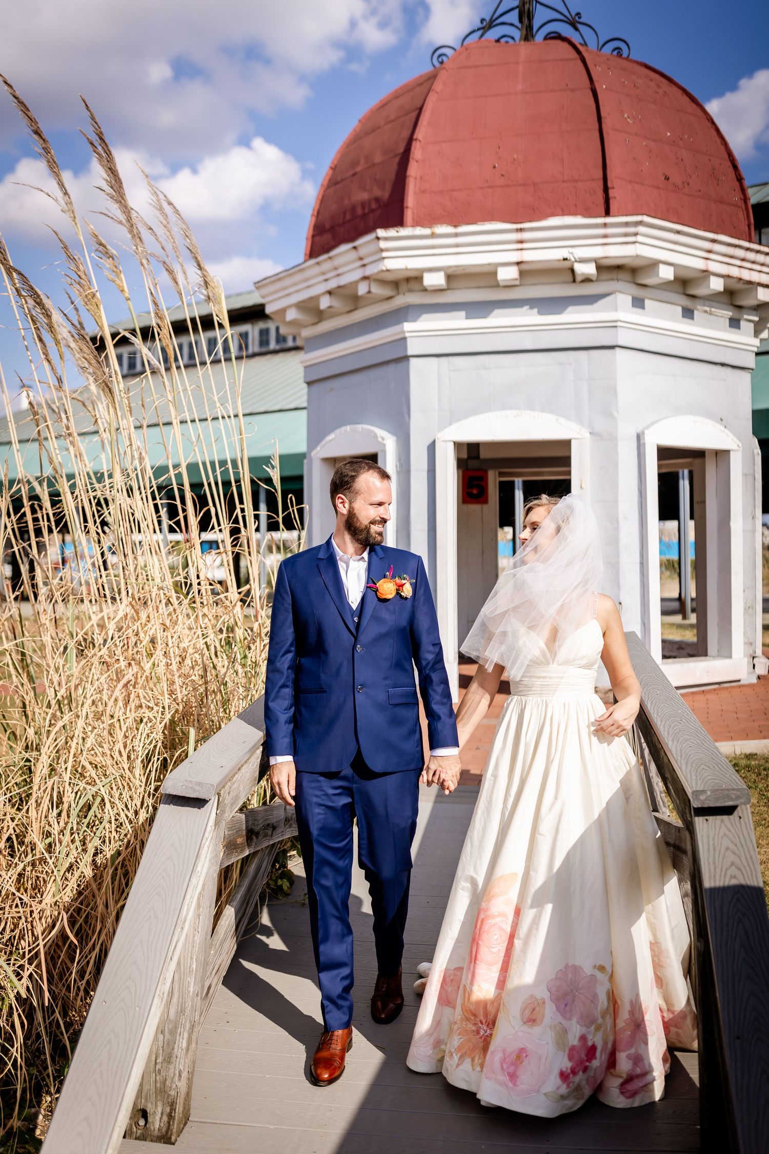 Couple in wedding attire walking on boardwalk near red-domed gazebo and coastal grass.