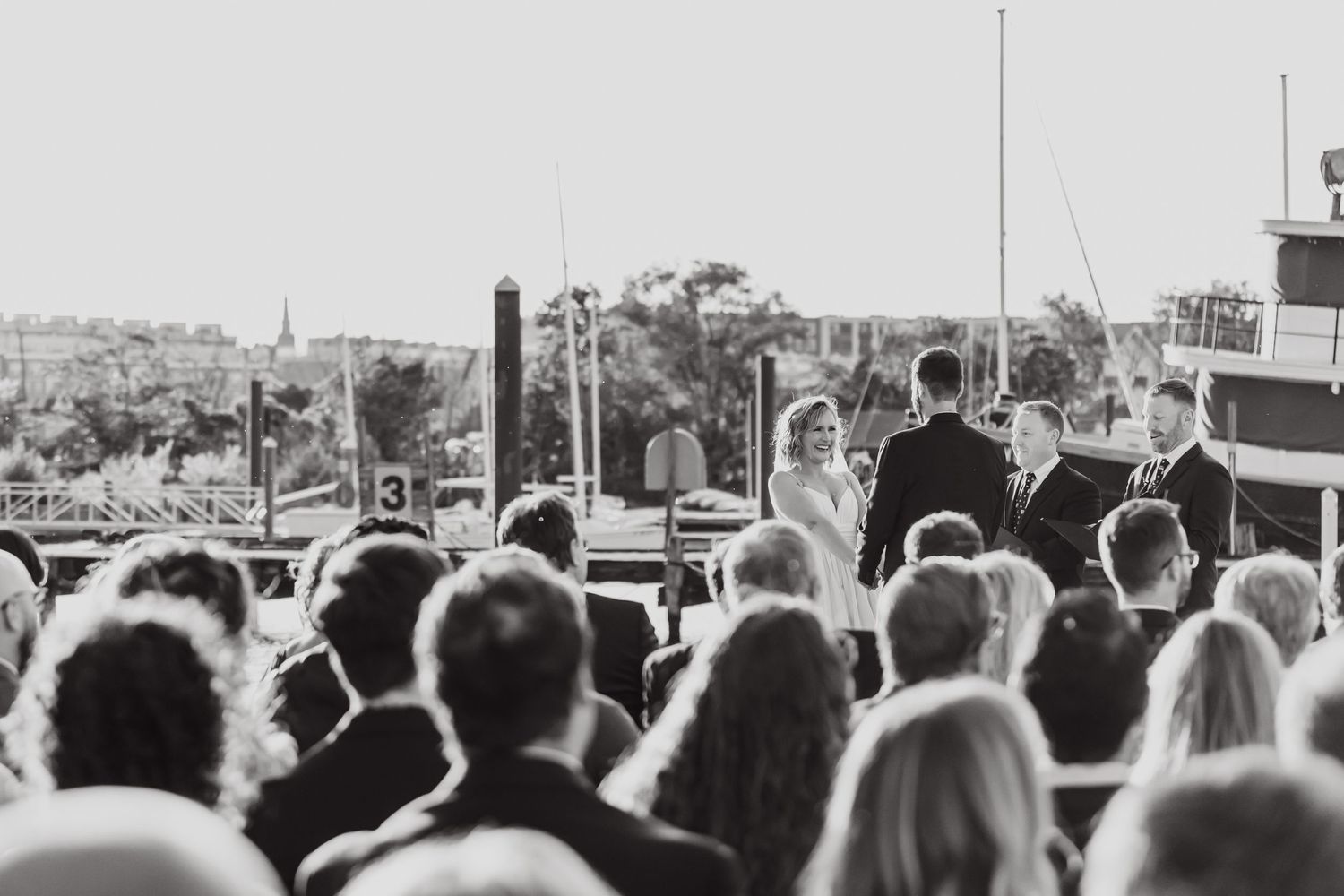 Black and white view of wedding ceremony crowd from behind facing waterfront.