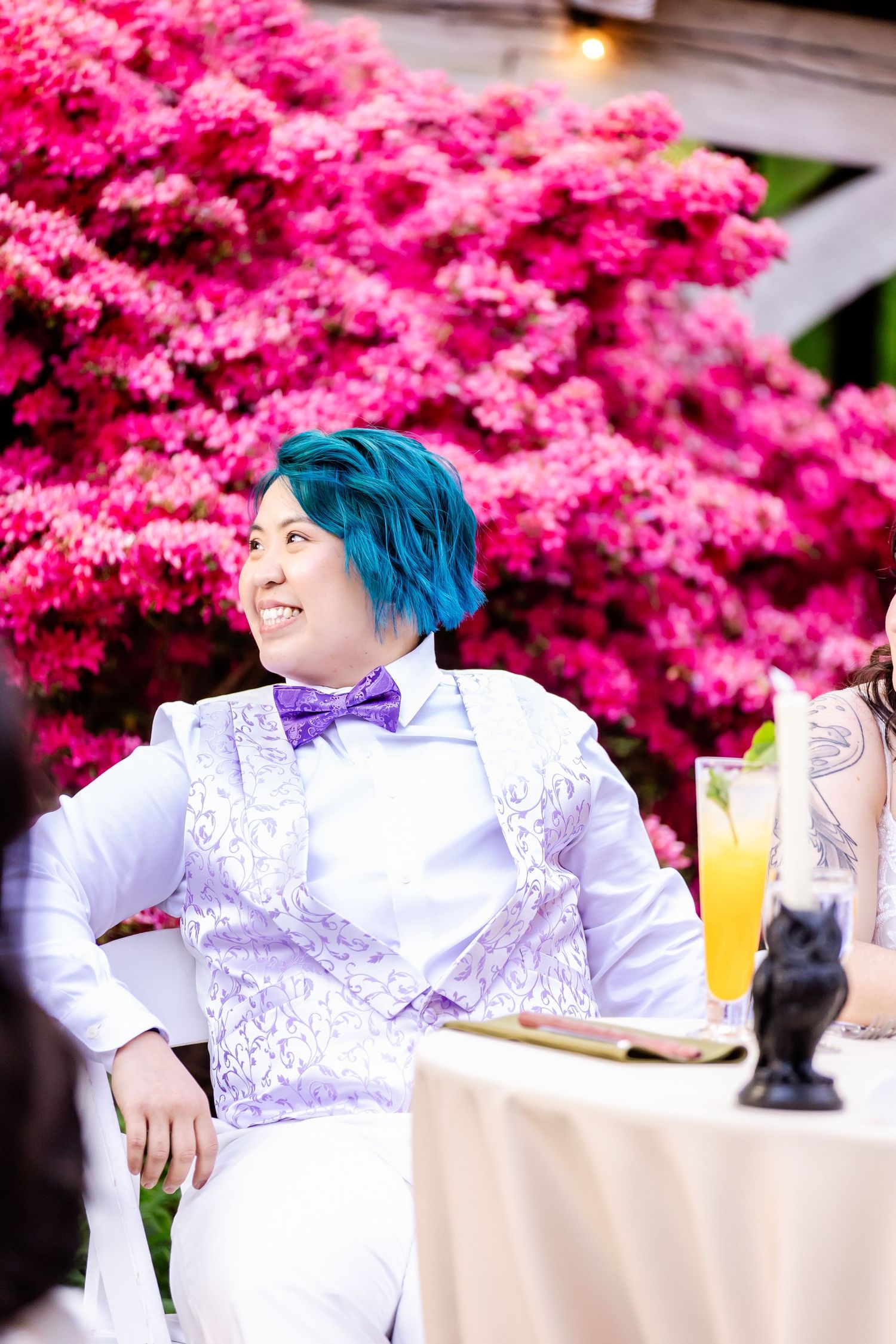 A person in white attire poses against vibrant pink azalea flowers at an outdoor event.