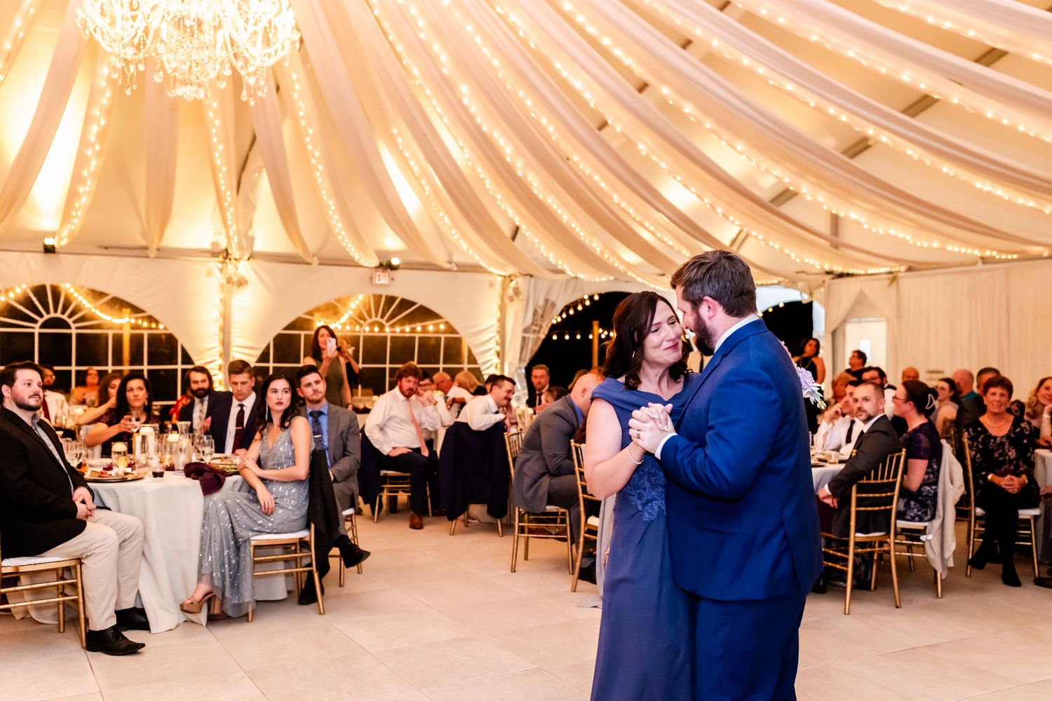 Couple dances intimately beneath draped fabric and chandeliers in an elegant wedding tent.