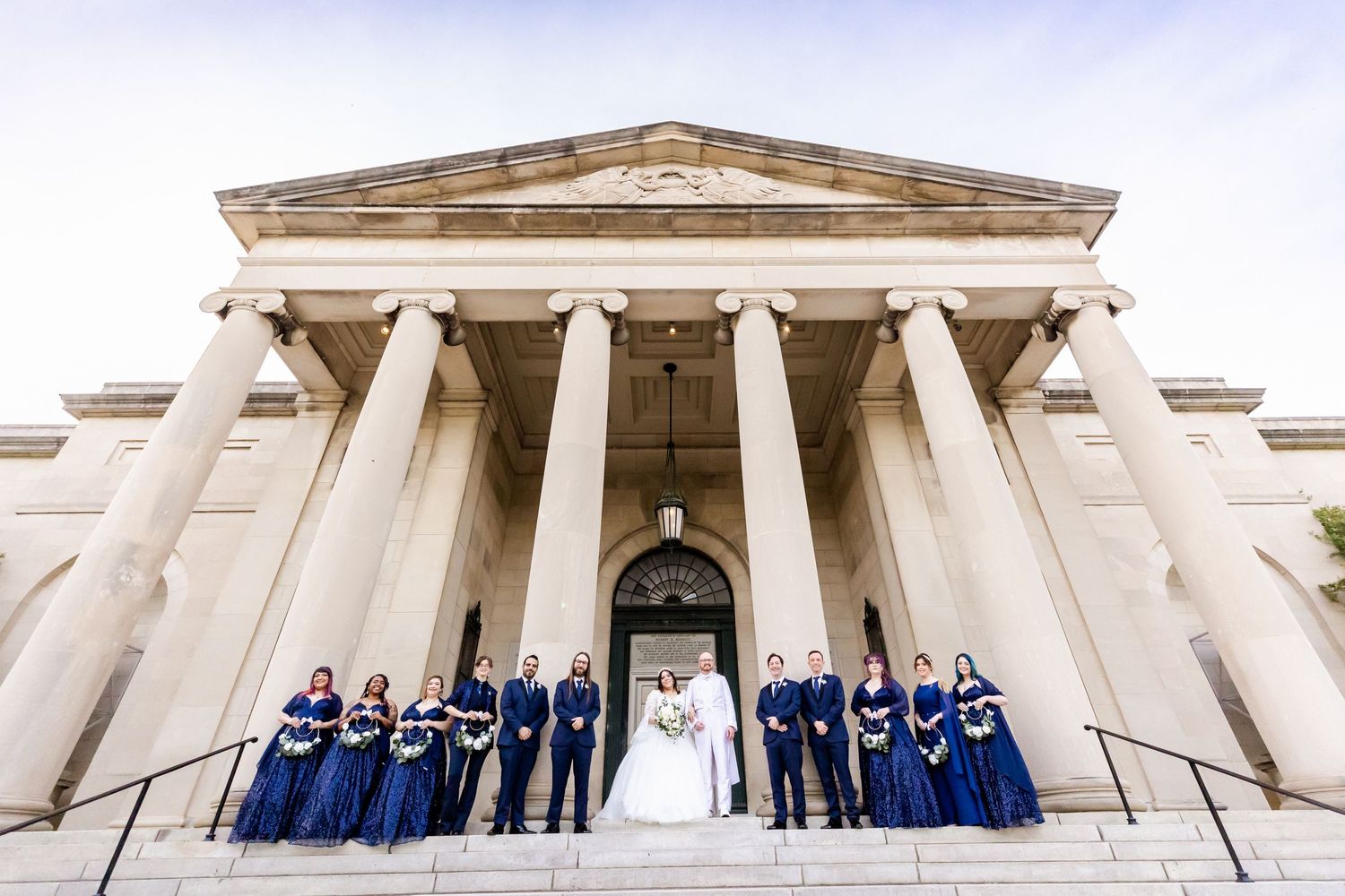 Wedding party in navy blue attire poses on grand marble steps of a neoclassical building with white columns.