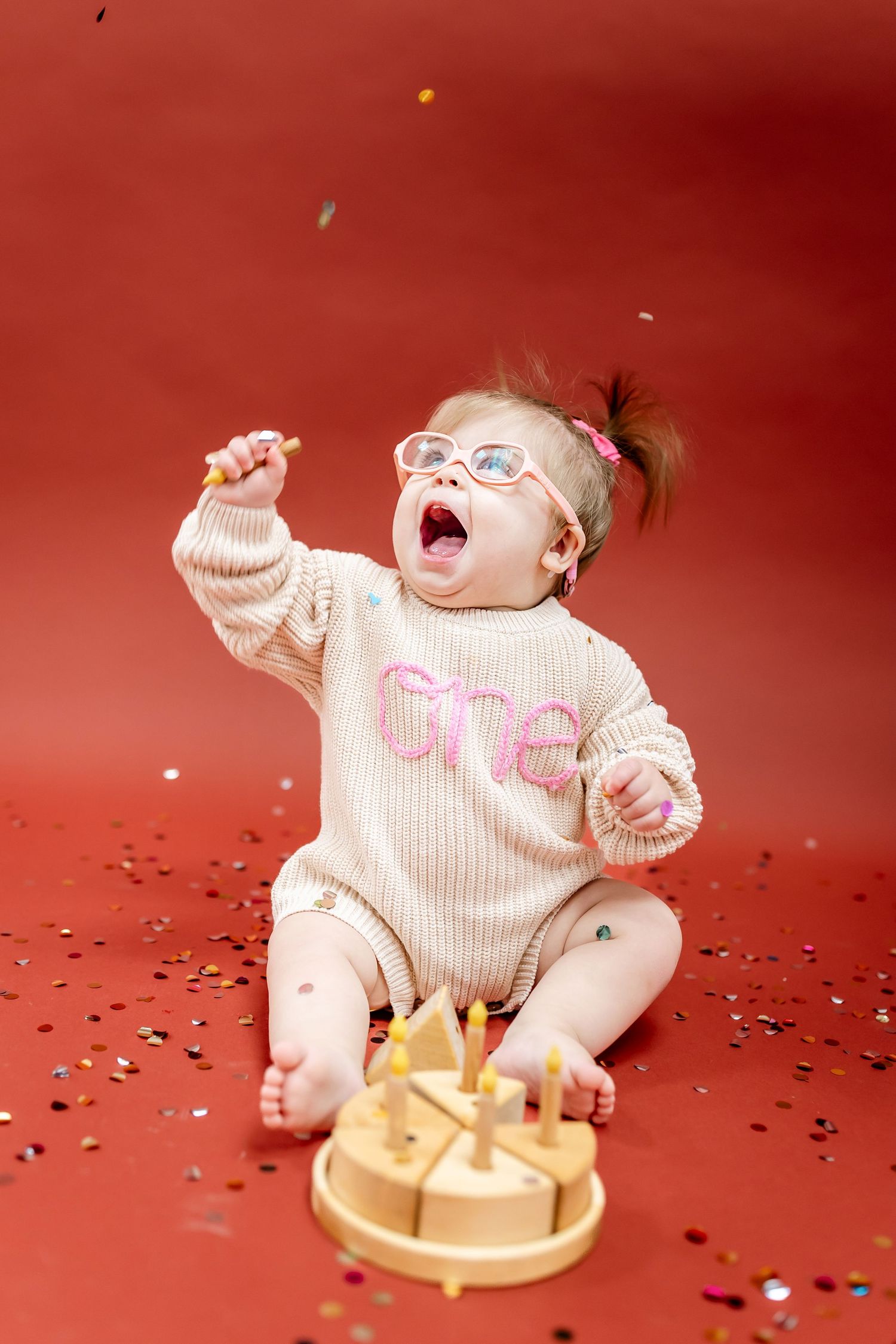 A baby in cream sweater reaches up with joy as confetti falls on red background.