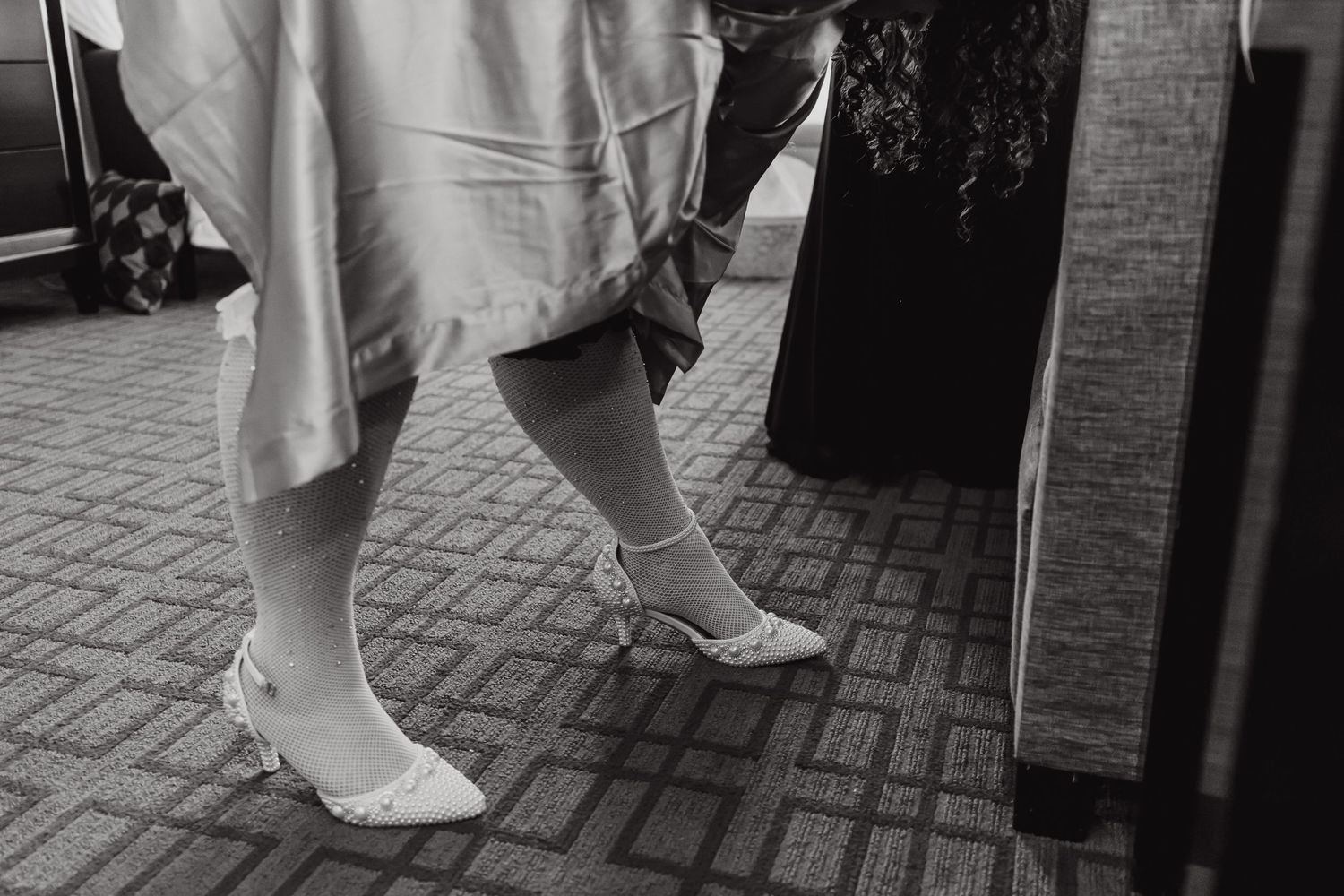 Close-up black and white photo of wedding shoes and dress hem on patterned carpet.
