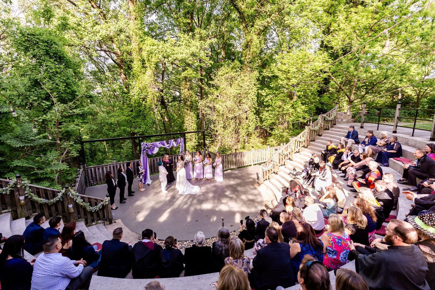 Aerial view of an outdoor wedding ceremony with guests seated on wooden benches in a forest setting.