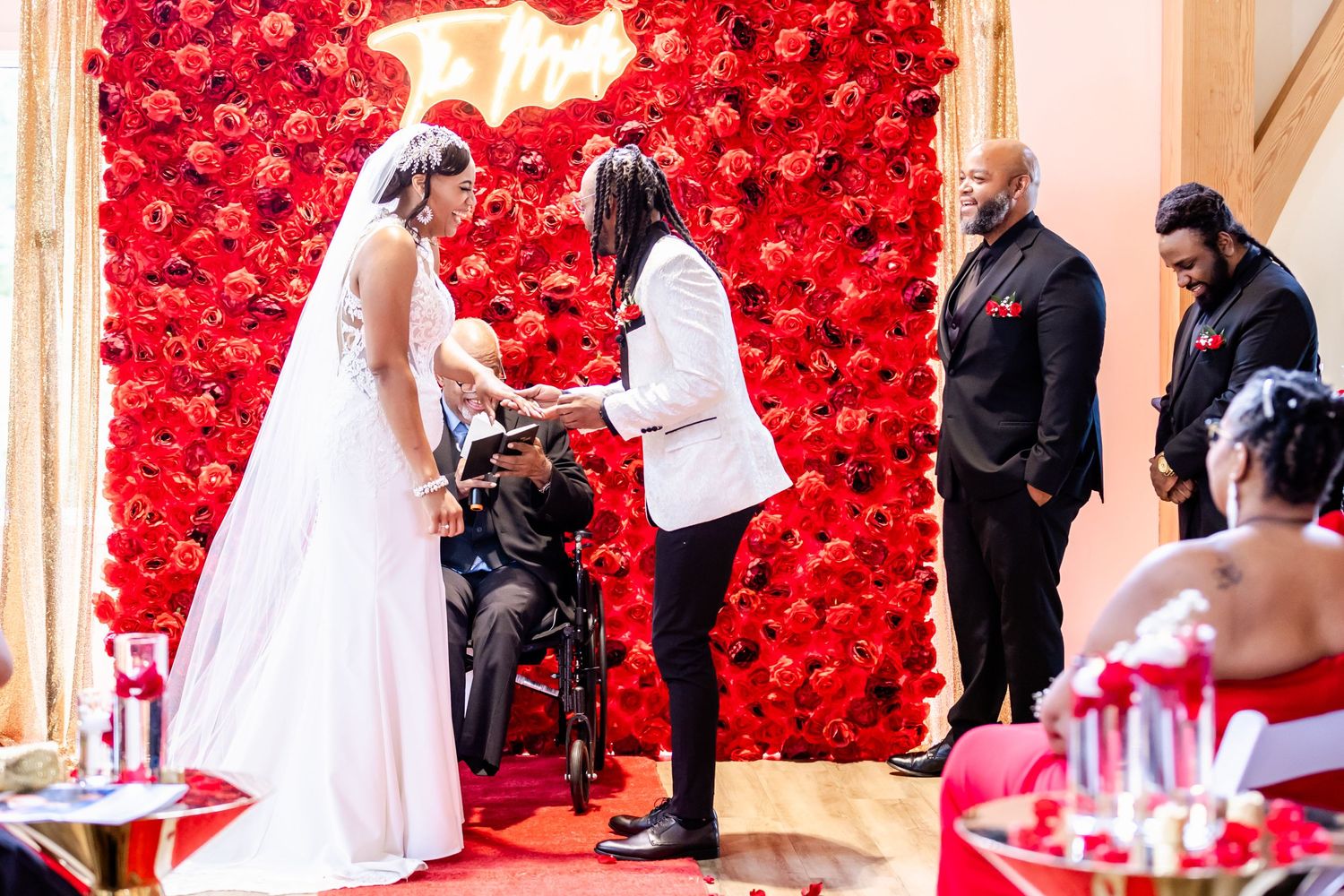 Wedding ceremony takes place in front of a stunning red rose wall backdrop during an intimate indoor celebration.