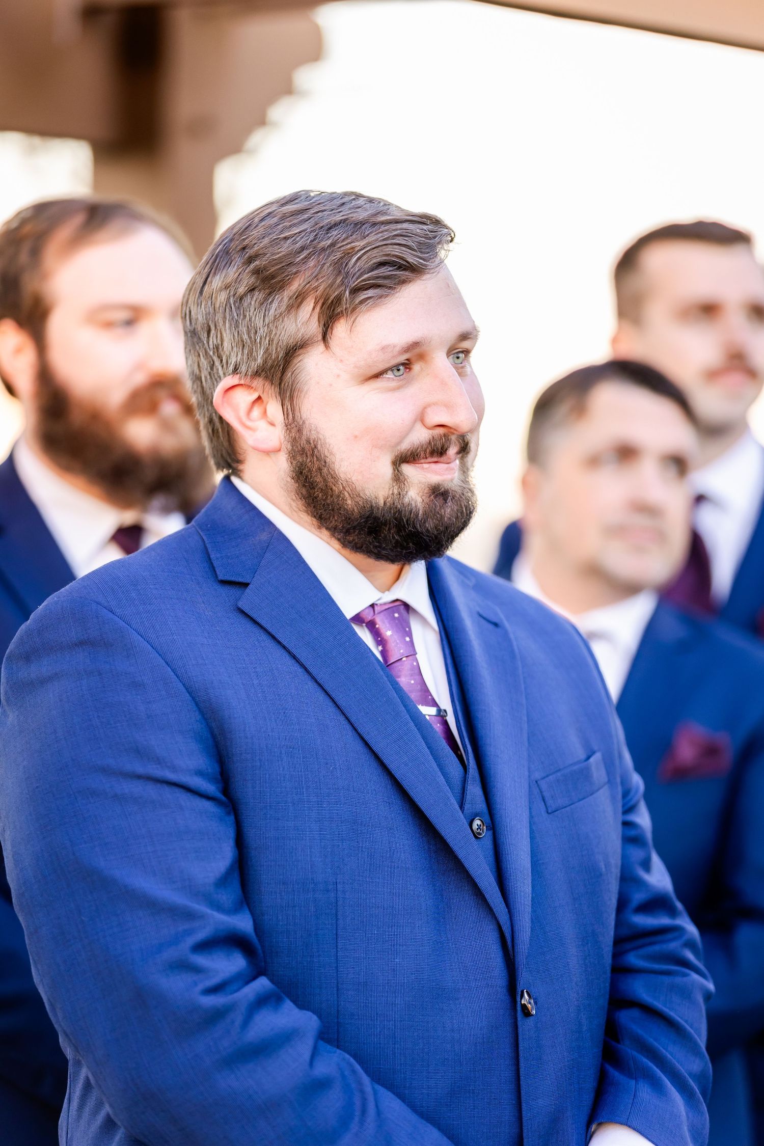 Groomsmen in formal attire watching wedding ceremony outdoors.