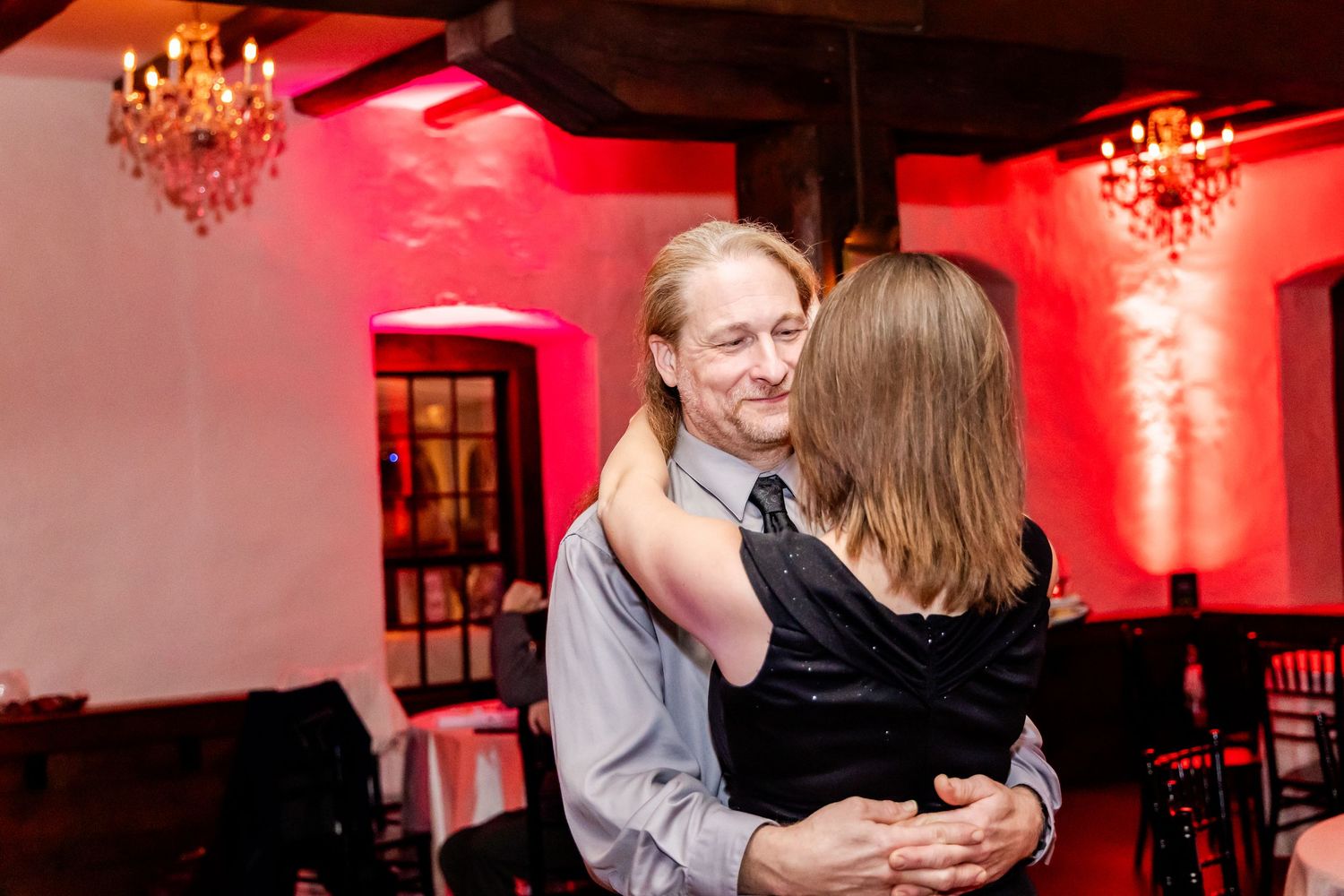A couple shares a romantic dance moment under red lighting with crystal chandeliers in an elegant venue.