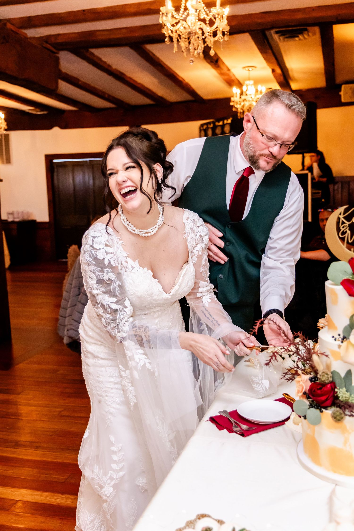 Wedding reception guests laughing and celebrating during cake cutting ceremony under elegant chandeliers.