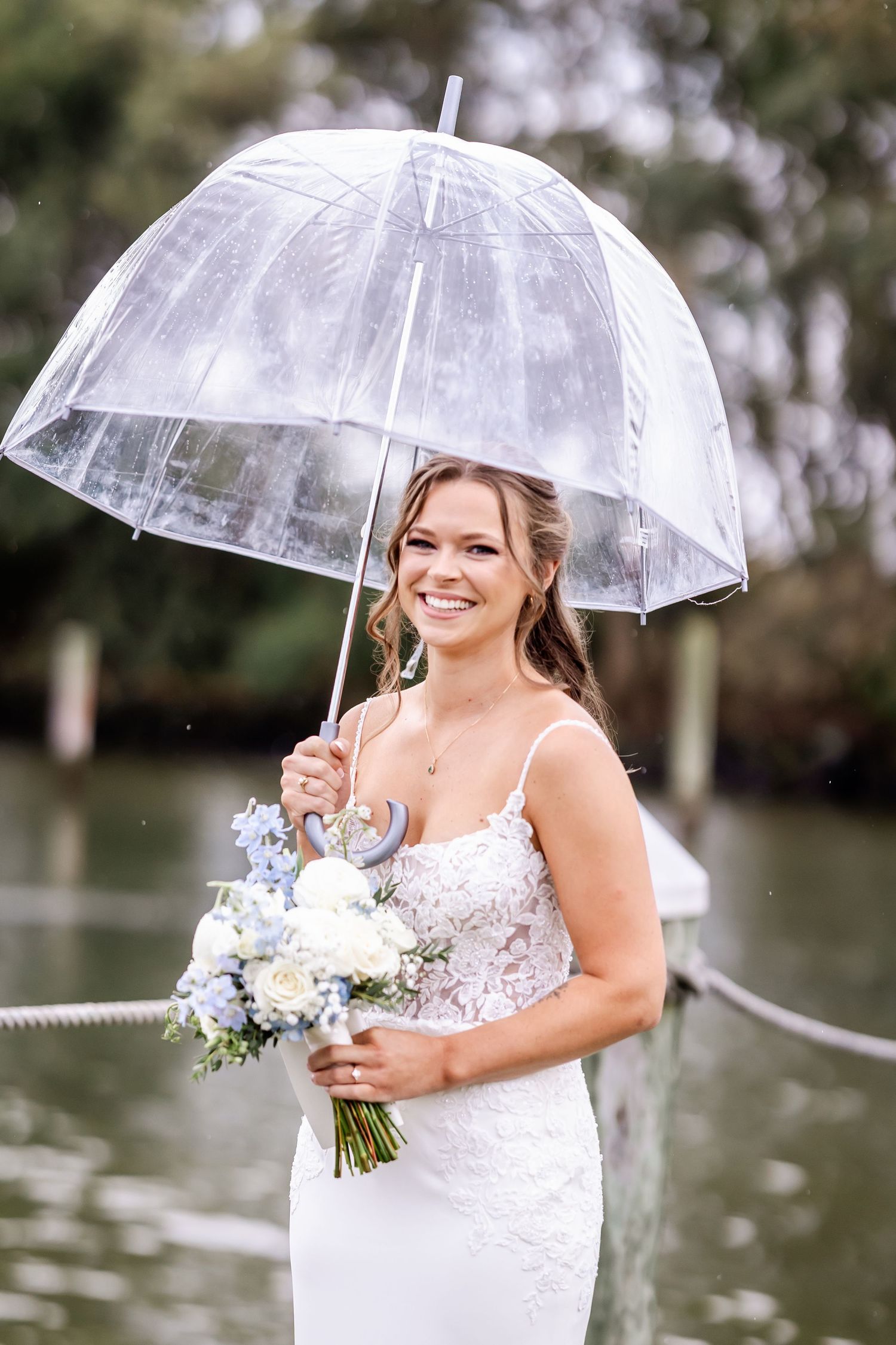 Bride in white dress holds clear umbrella and bouquet while smiling during rainy wedding photoshoot.