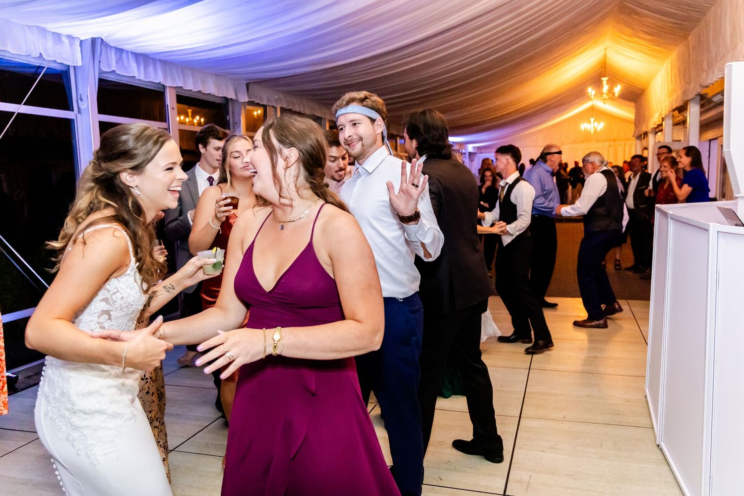 Wedding reception guests dancing and celebrating in an elegant white tent with purple lighting.