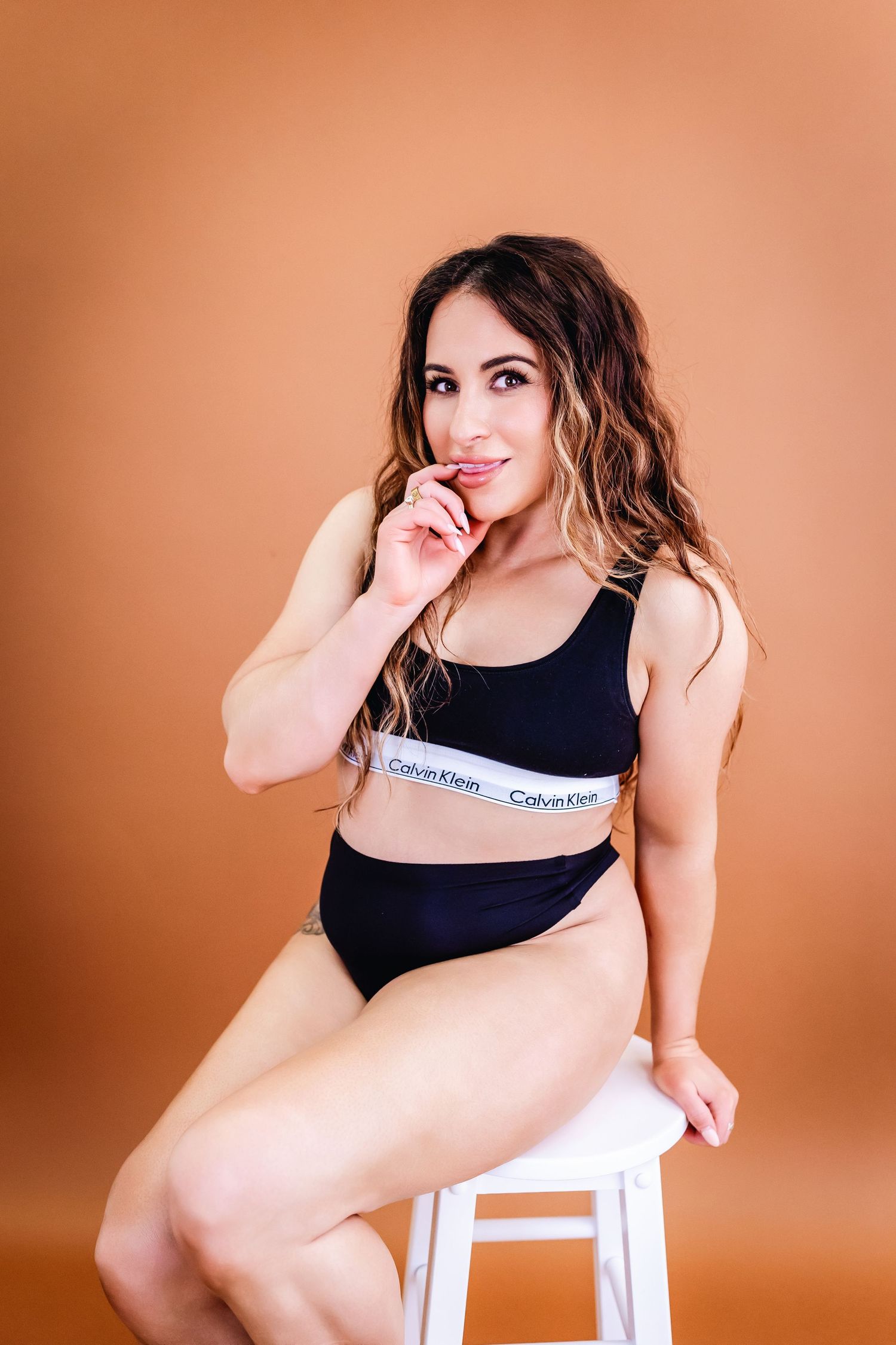 Model in black Calvin Klein sports bra poses against peach backdrop in artistic boudoir photoshoot series.