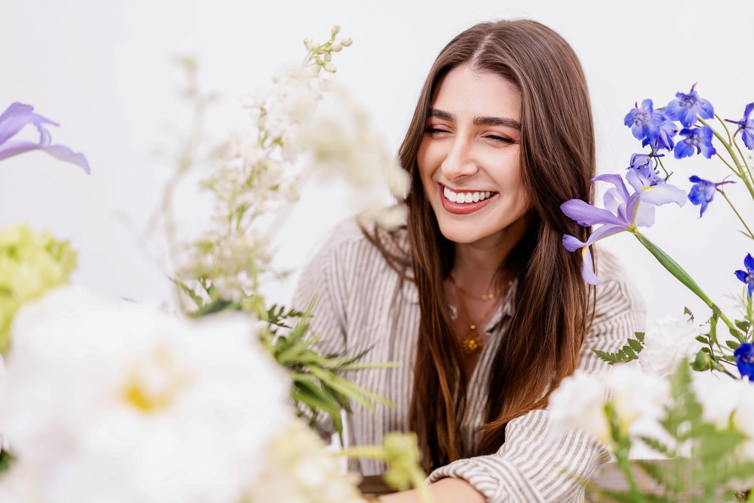 Smiling woman with long brown hair laughing among fresh white and purple flowers in a bright setting.