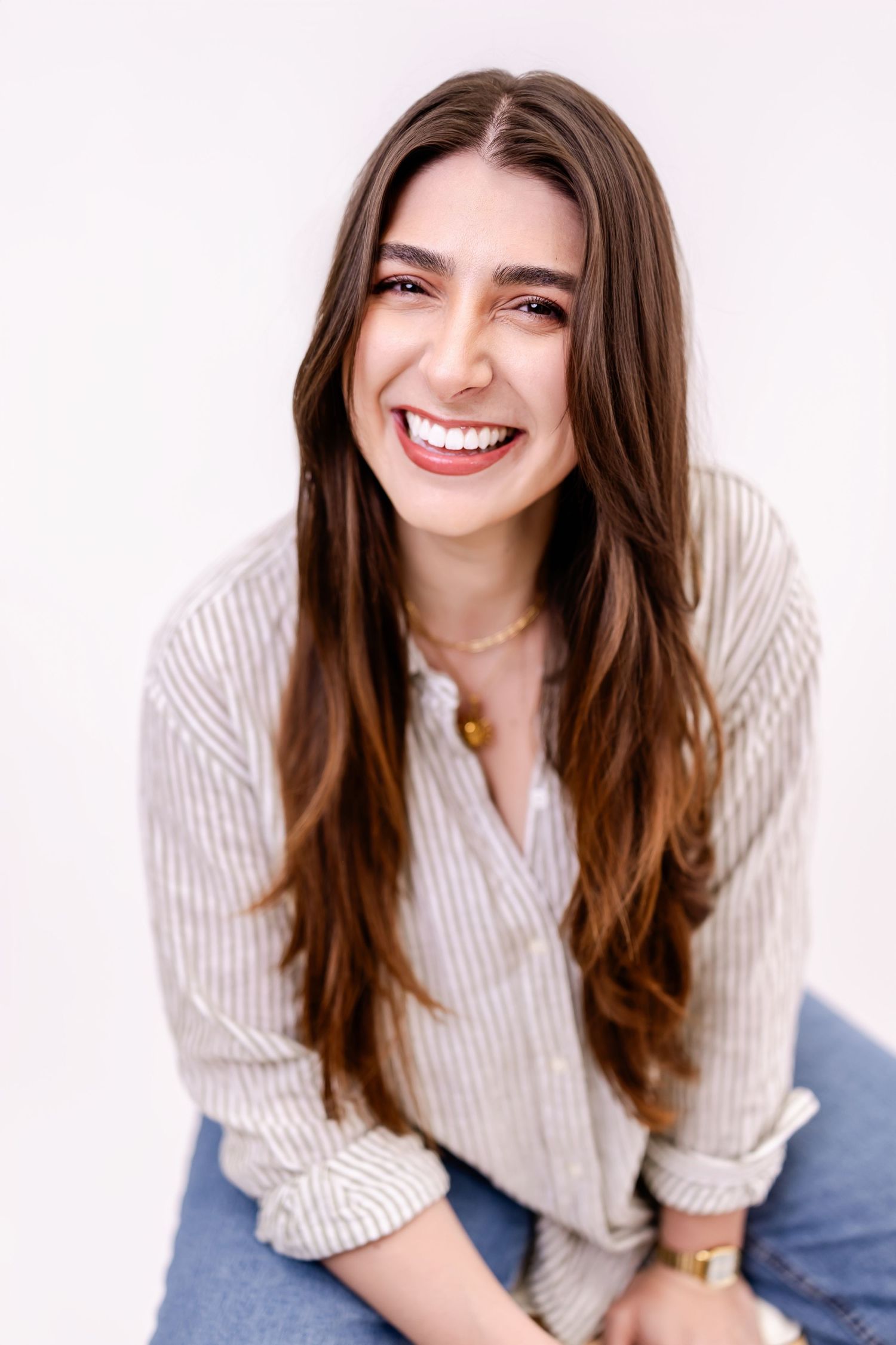Smiling young woman with long brown hair wearing a striped shirt and jeans, posing against a white background.