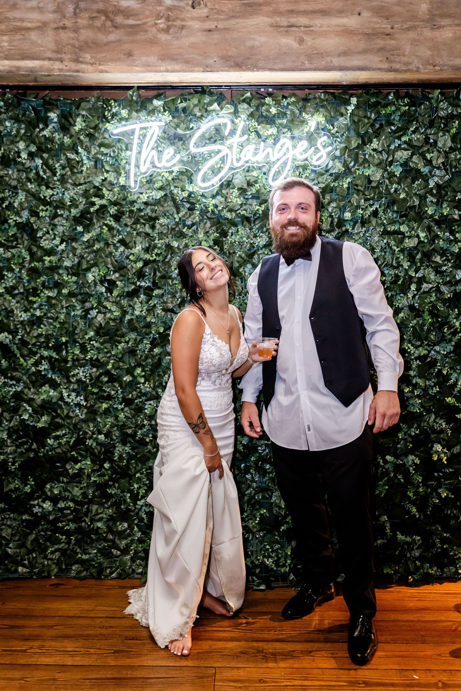 Newlyweds pose in front of a green ivy wall with a neon sign reading The Stangers at their wedding reception.