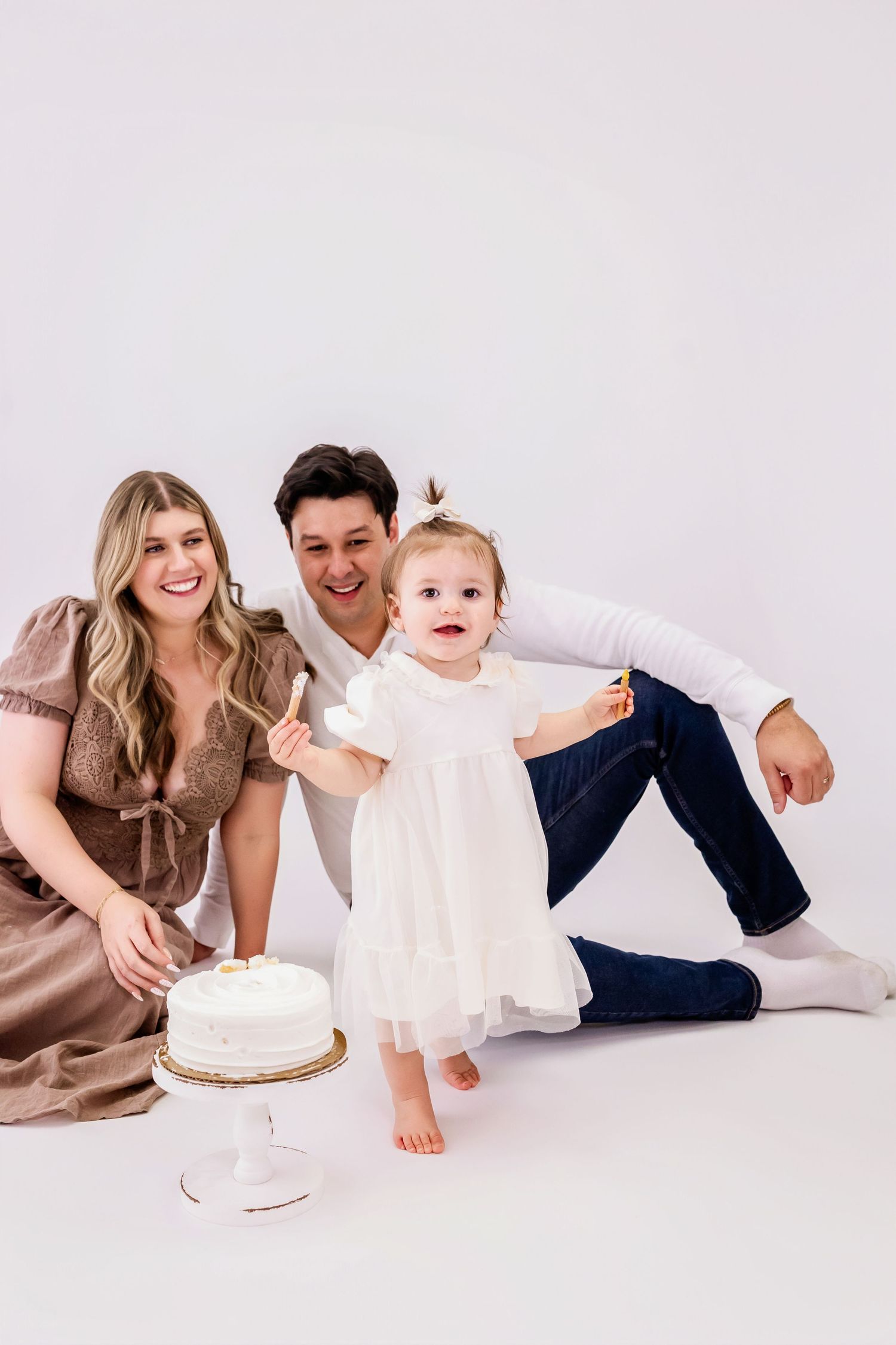 A family in white and beige outfits lies on the floor together in a minimalist studio setting for a portrait series.
