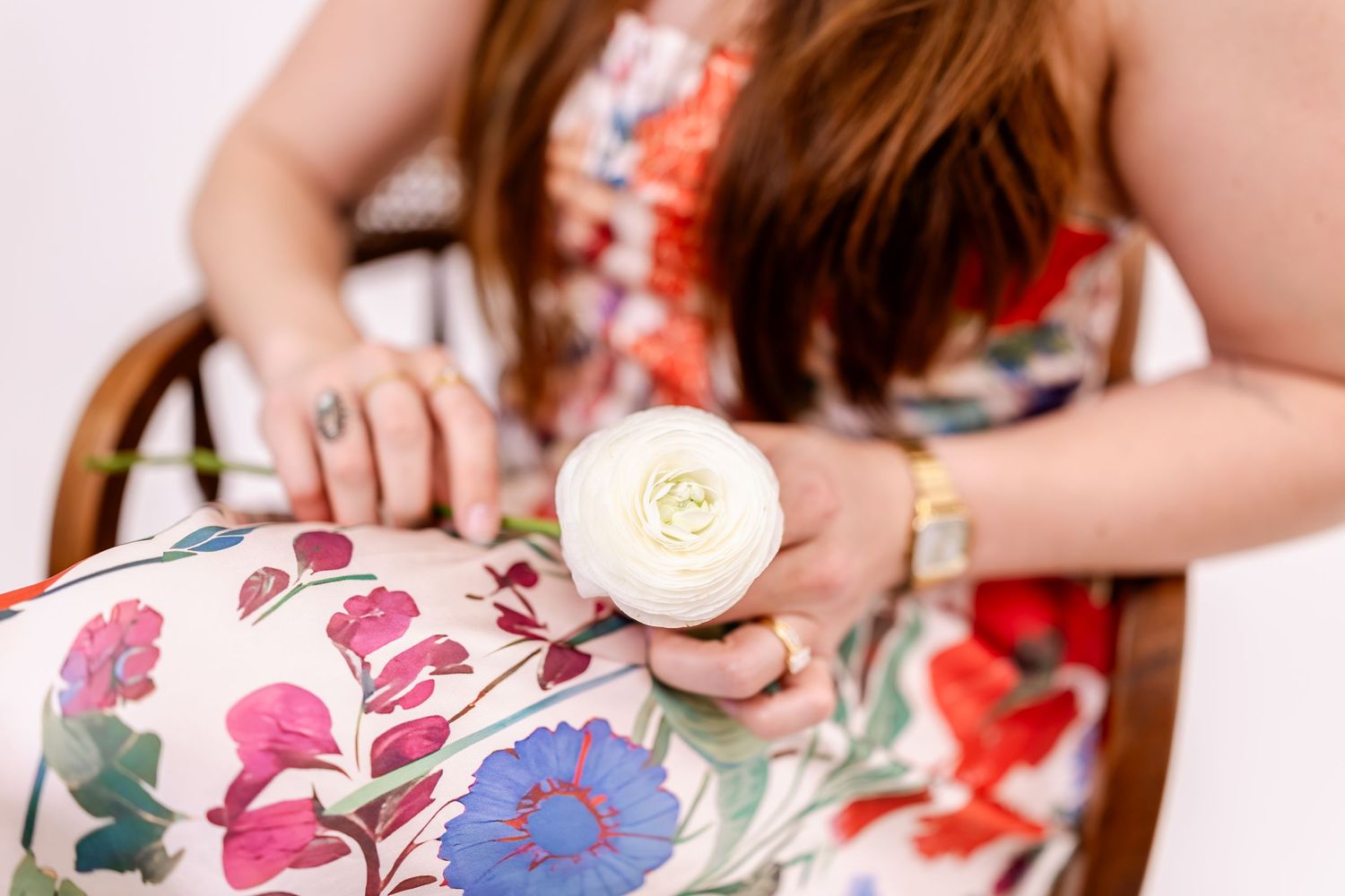 Woman in floral dress holding a white ranunculus flower while seated in a colorful floral chair.
