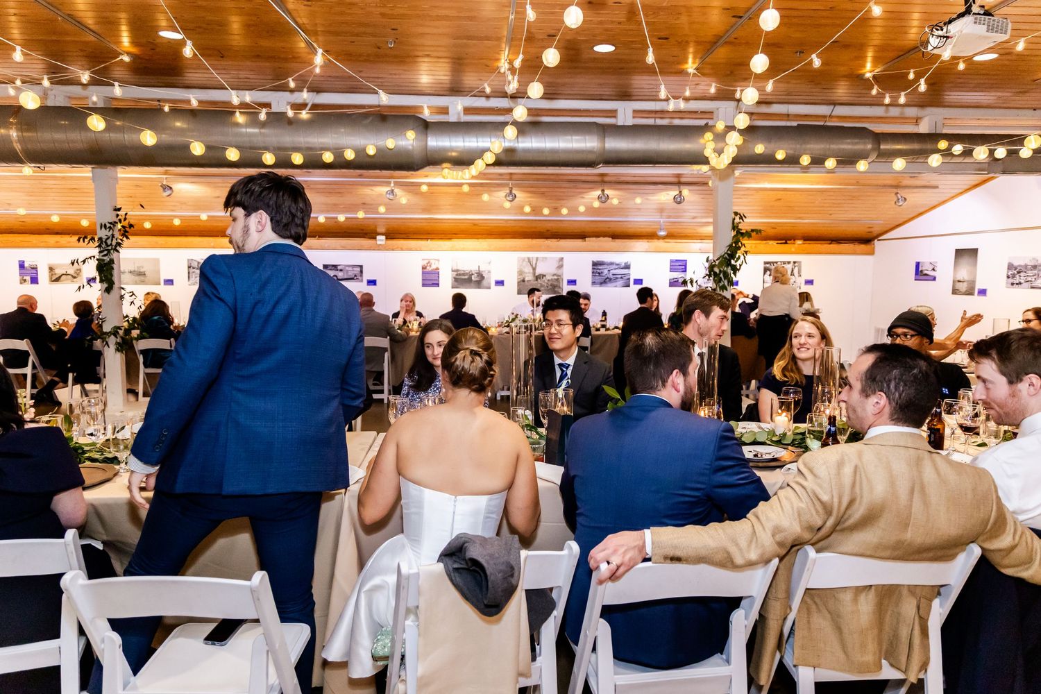 Elegant wedding reception in a warmly lit venue with string lights, guests seated at tables, bride and groom at center.