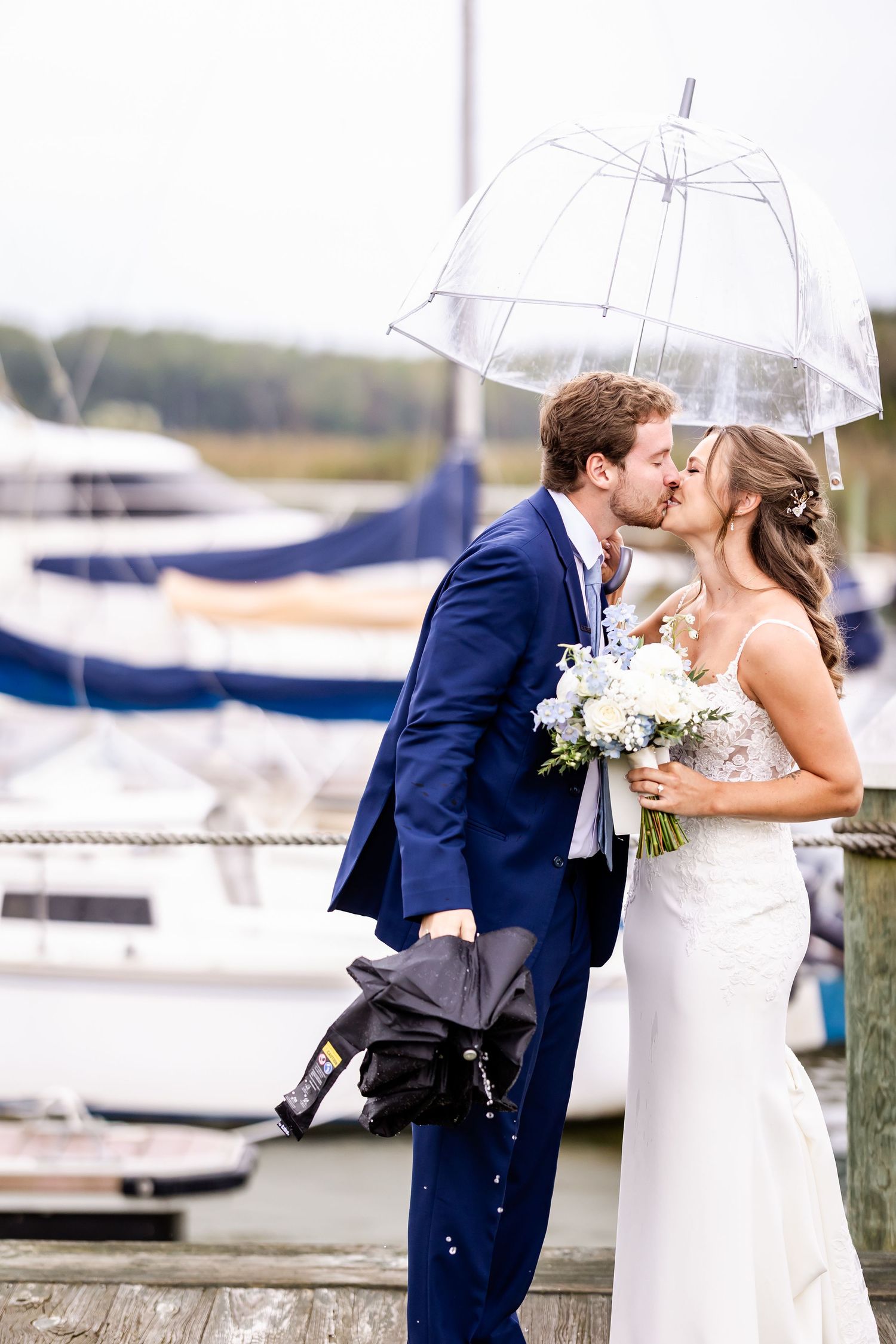 Romantic wedding couple shares a kiss by a marina with boats in the background on an overcast day.