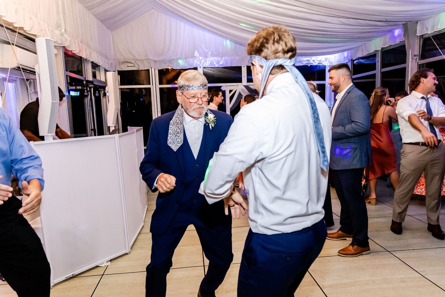 Wedding guests dance and celebrate in a white draped tent venue with festive lighting.