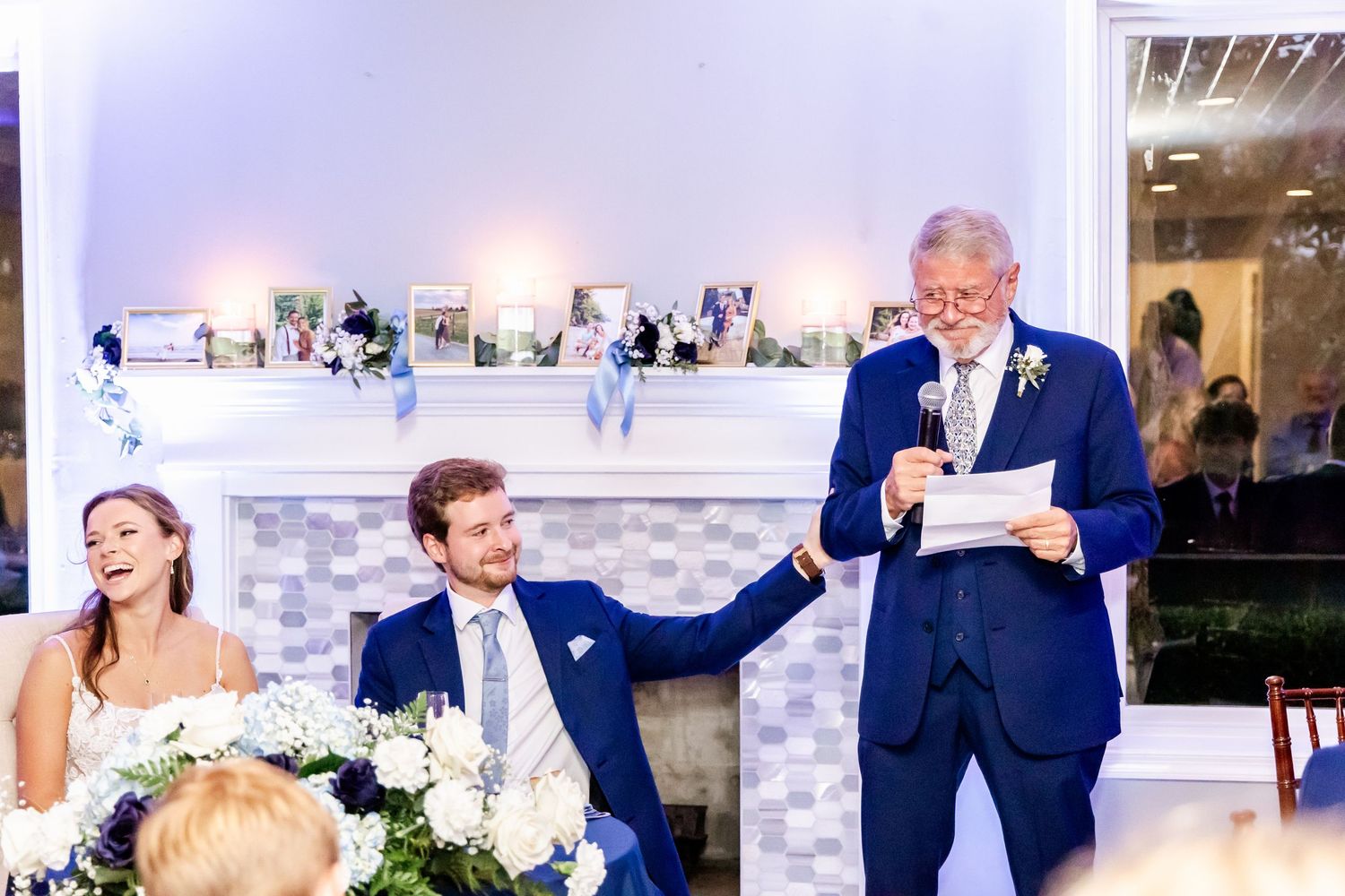 Wedding reception speech at an elegant venue with white mantle and decorative photo display.