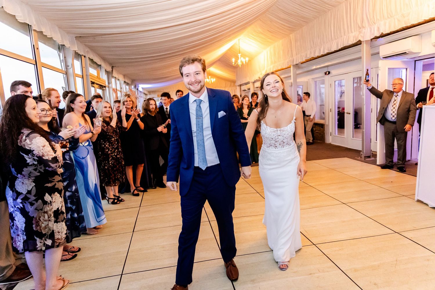 Wedding guests dancing and celebrating on a brightly lit dance floor under draped white fabric ceiling.
