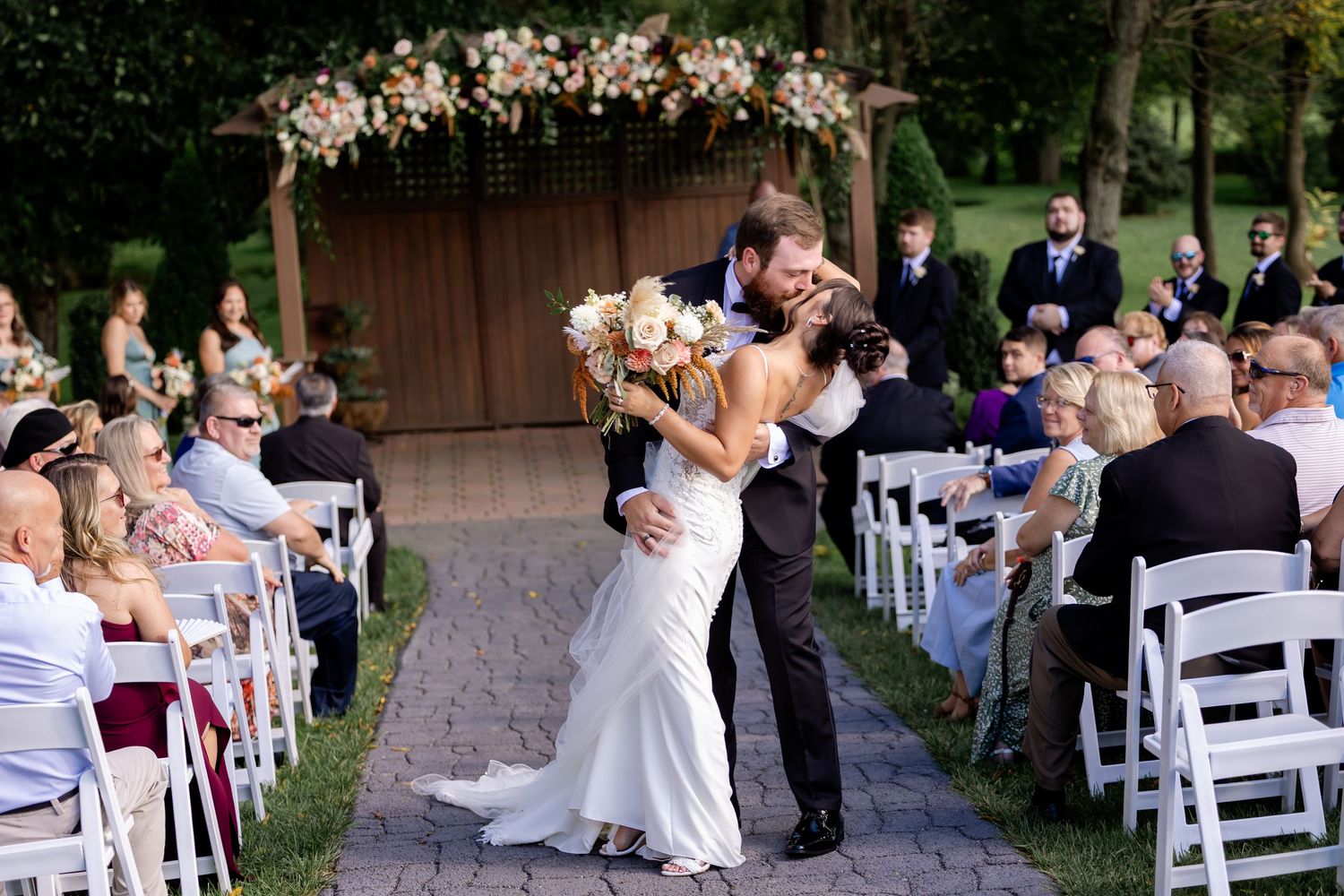 Bride and groom share a romantic kiss at the altar during an outdoor wedding ceremony with white chairs and floral arch.