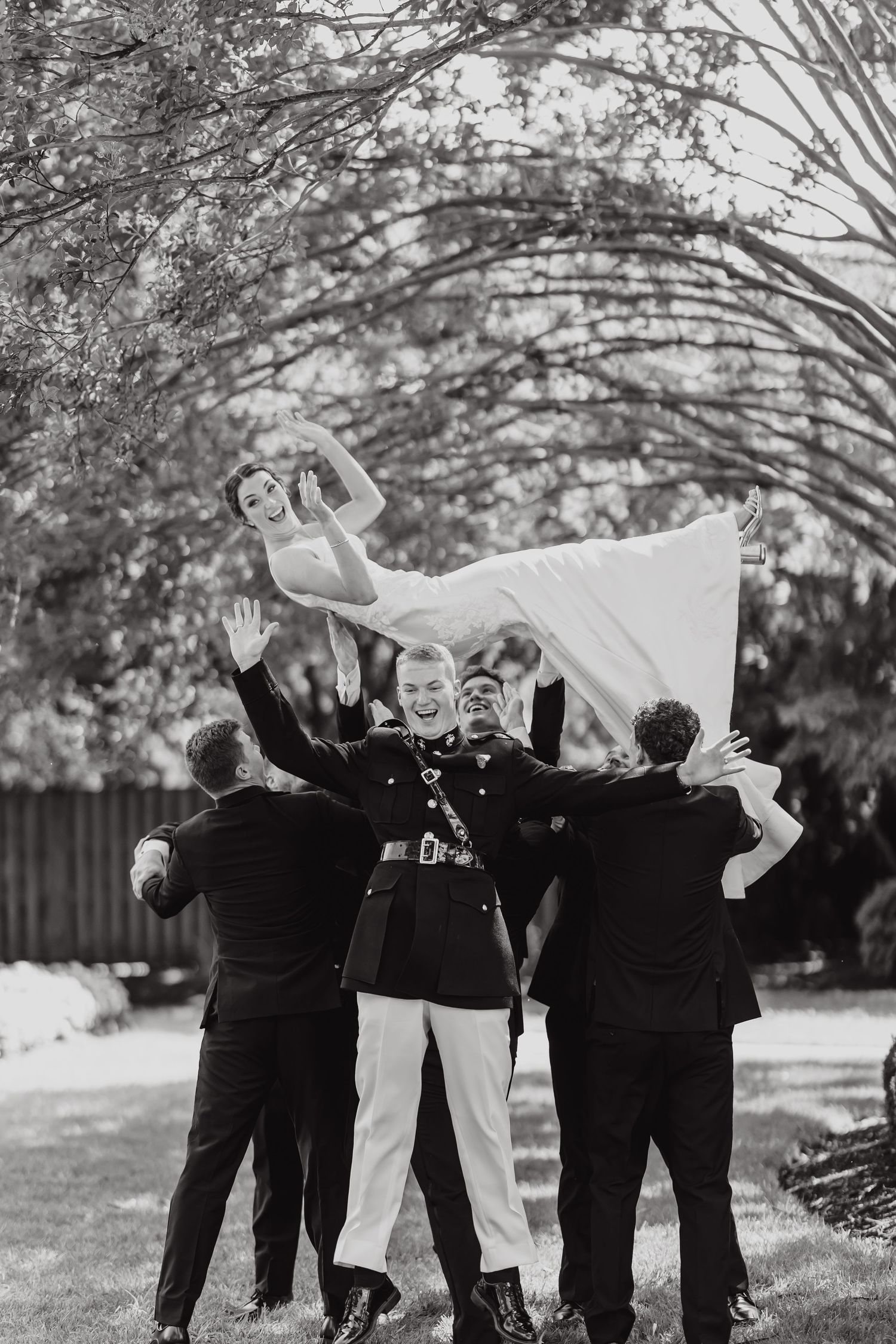 Black and white wedding photo showing a joyful group tossing someone in a flowing white dress in an outdoor garden setting.