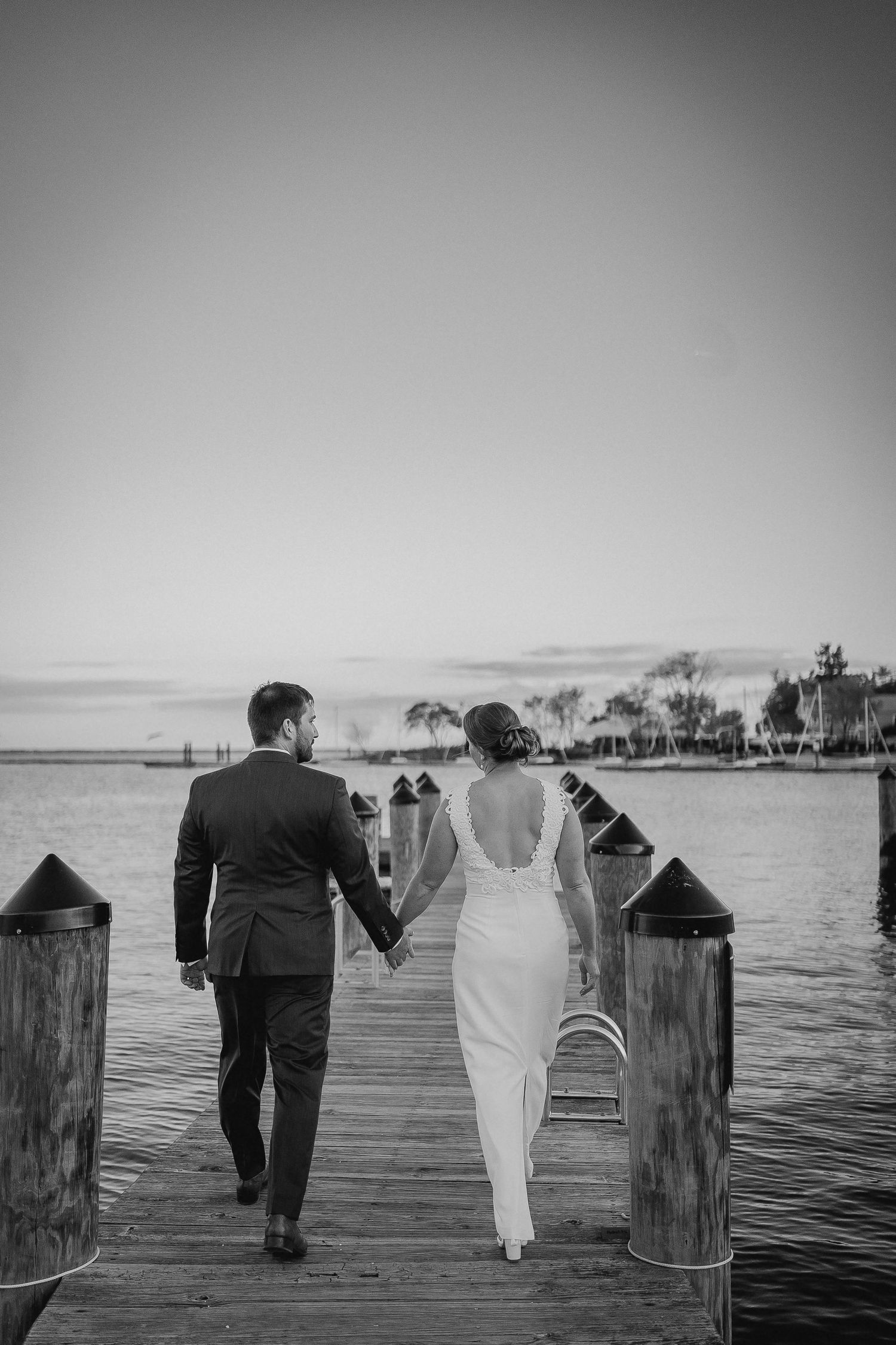 A couple walks along a wooden pier over water in black and white, with silhouettes against a bright horizon.